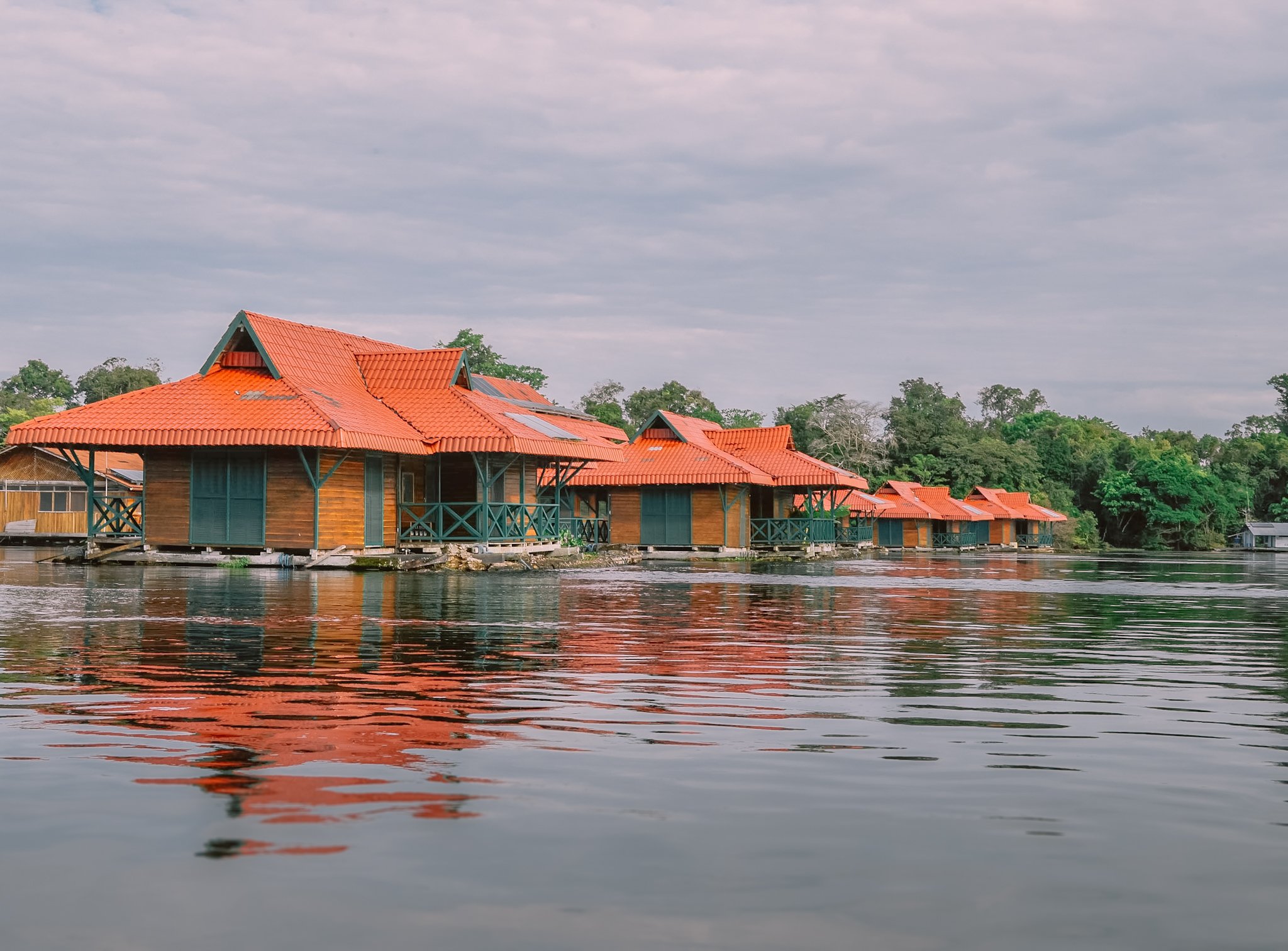 Orange rooftops doubling in the still river — the Uakari Lodge from the water. A row of wooden overwater cabins with vivid orange-tiled roofs reflected in calm river water, part of an amazon rainforest ecolodge trip in brazil surrounded by dense tropical forest