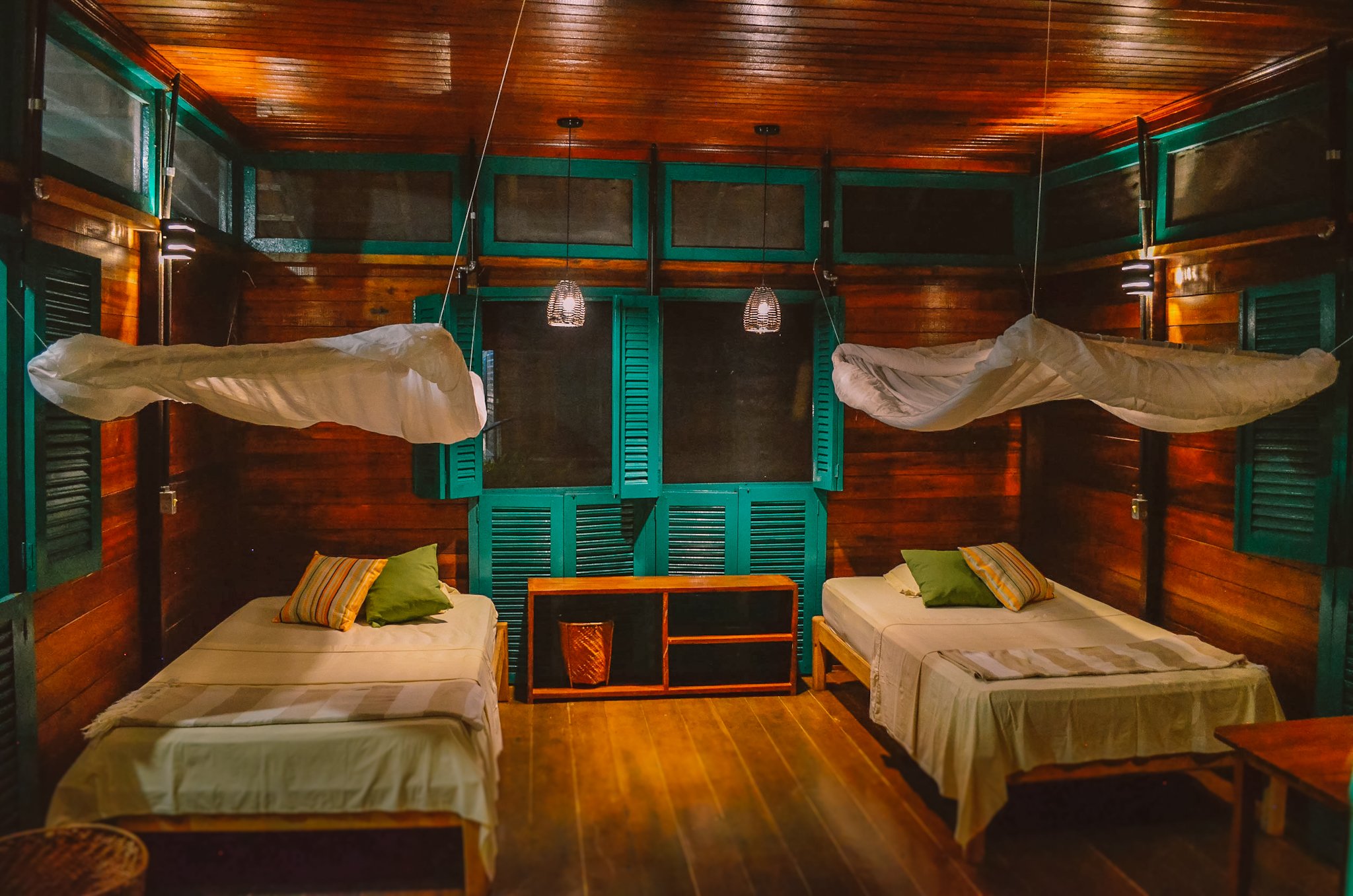 Zweibettzimmer in einem Amazonas-Ökolodge in Brasilien mit aufgehängten Moskitonetzen, warmem Holzvertäfelung und türkisfarbenen Fensterläden