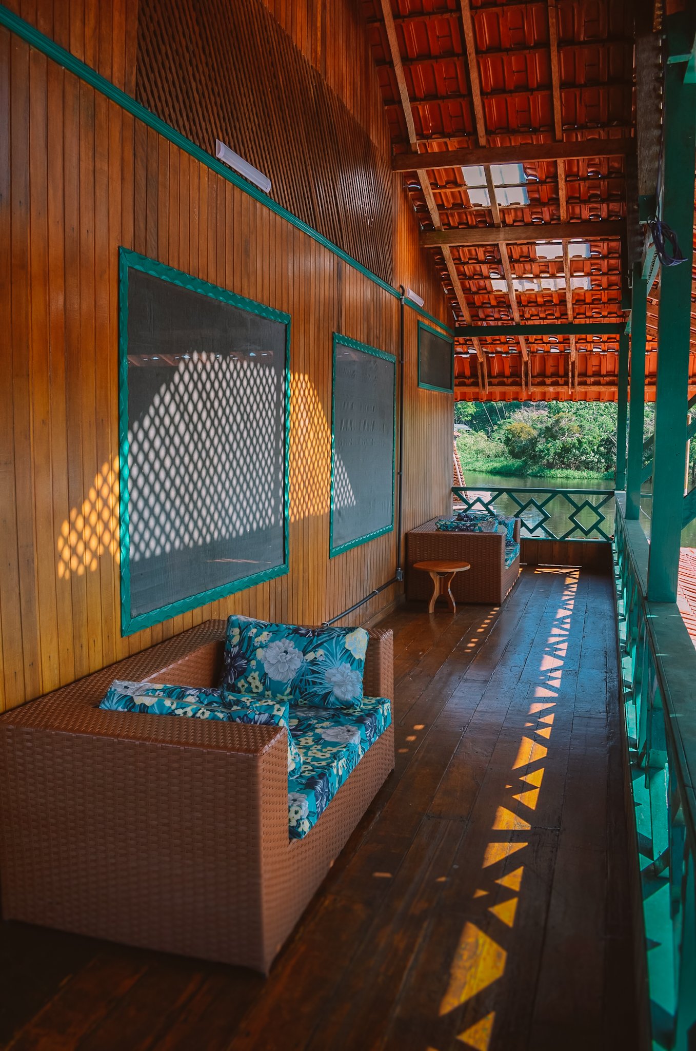 Überdachte Holzveranda einer Amazonia-Ecolodge mit Korbsesseln, tropischen Kissen und Sonnenlicht, das Gitterschatten auf den Holzdielen wirft, mit Blick auf eine grüne Flusslandschaft
