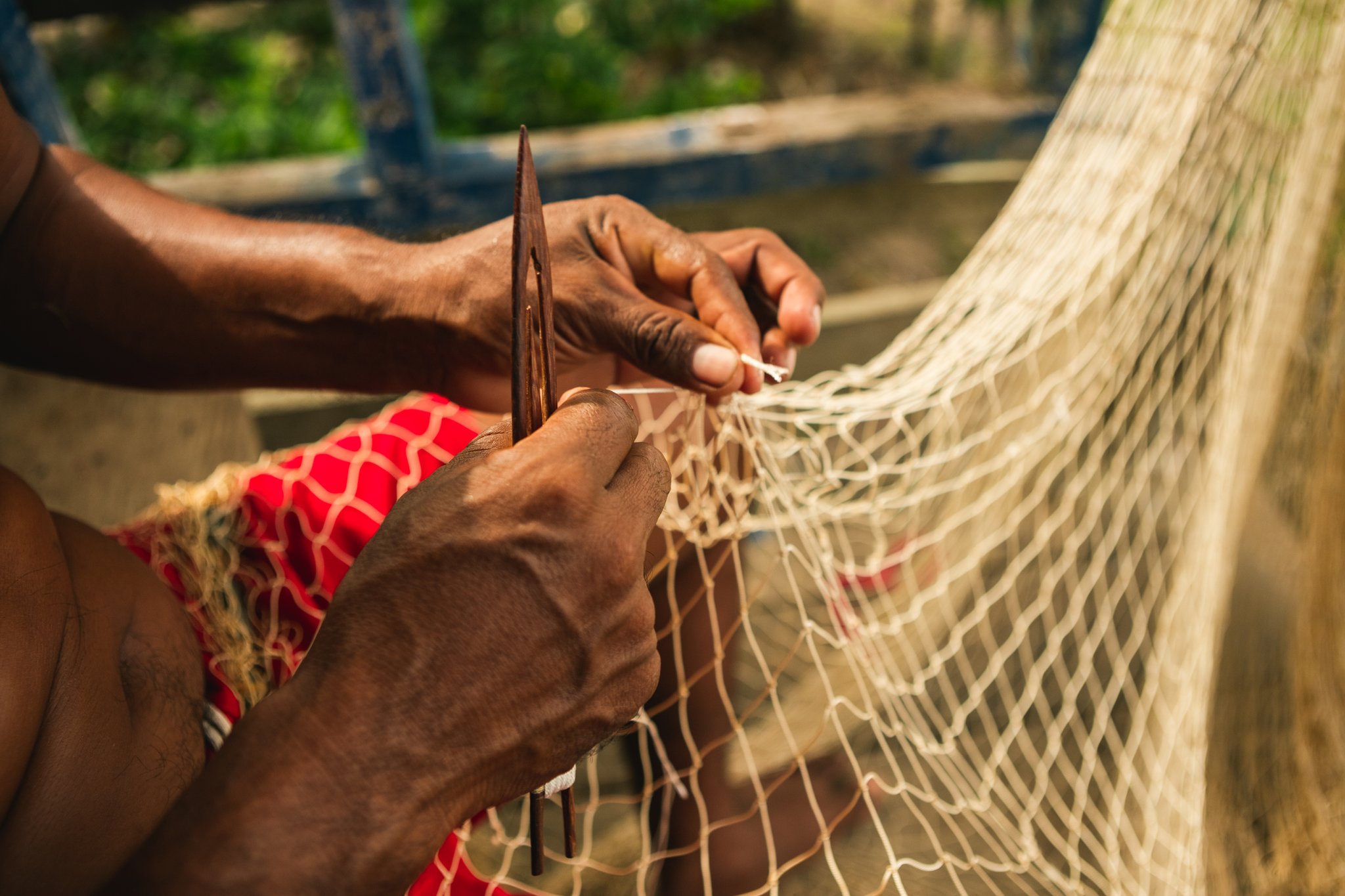 Knot by knot, a tradition passed down through riverside generations. Photo: Guilherme Gomes Close-up of skilled hands using a wooden needle to mend a fishing net during an amazon rainforest ecolodge trip in brazil, with green vegetation softly blurred in the background