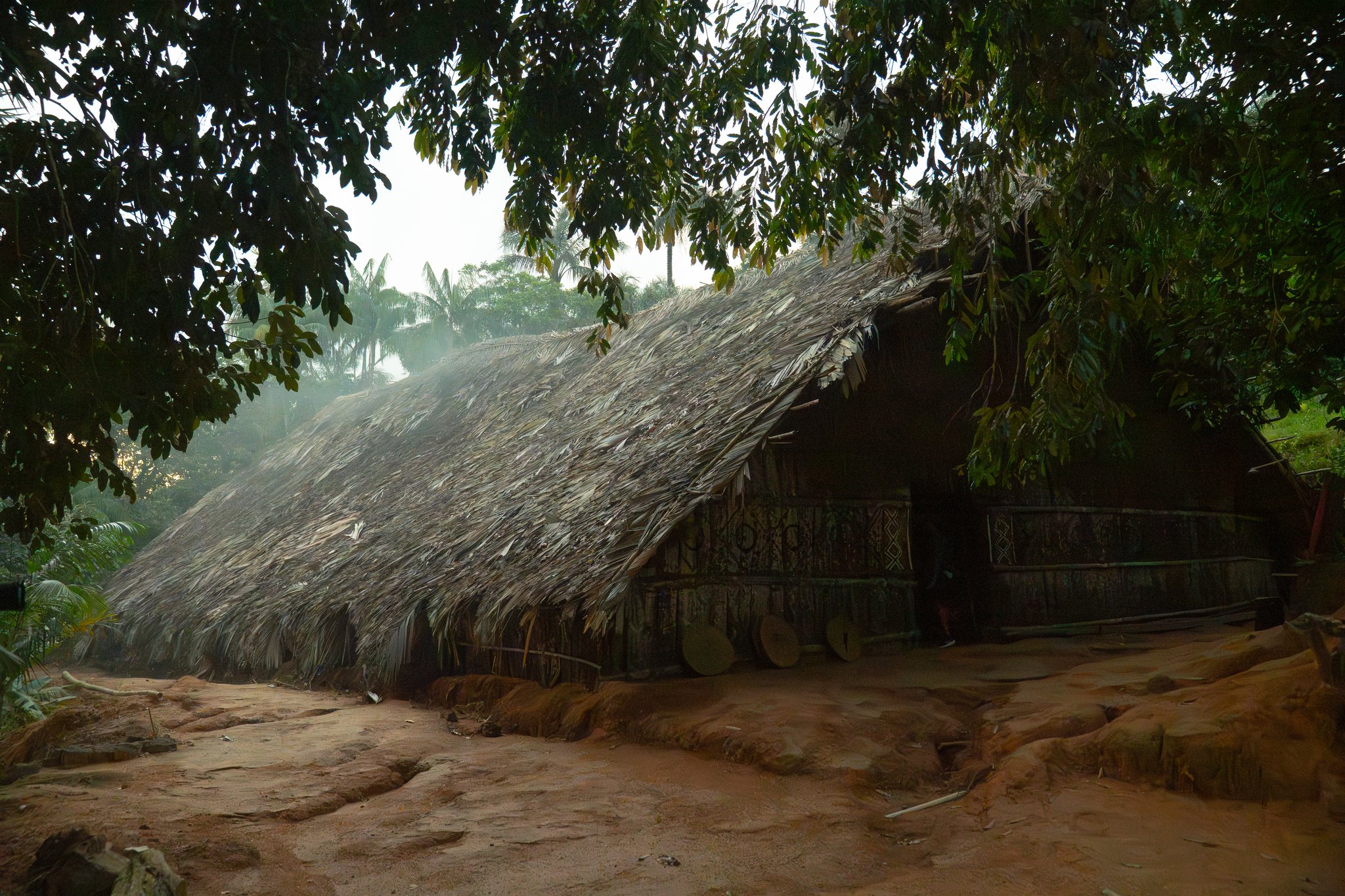 Traditional thatched-roof indigenous longhouse surrounded by misty Amazon rainforest trees and red earth, decorated with tribal geometric patterns