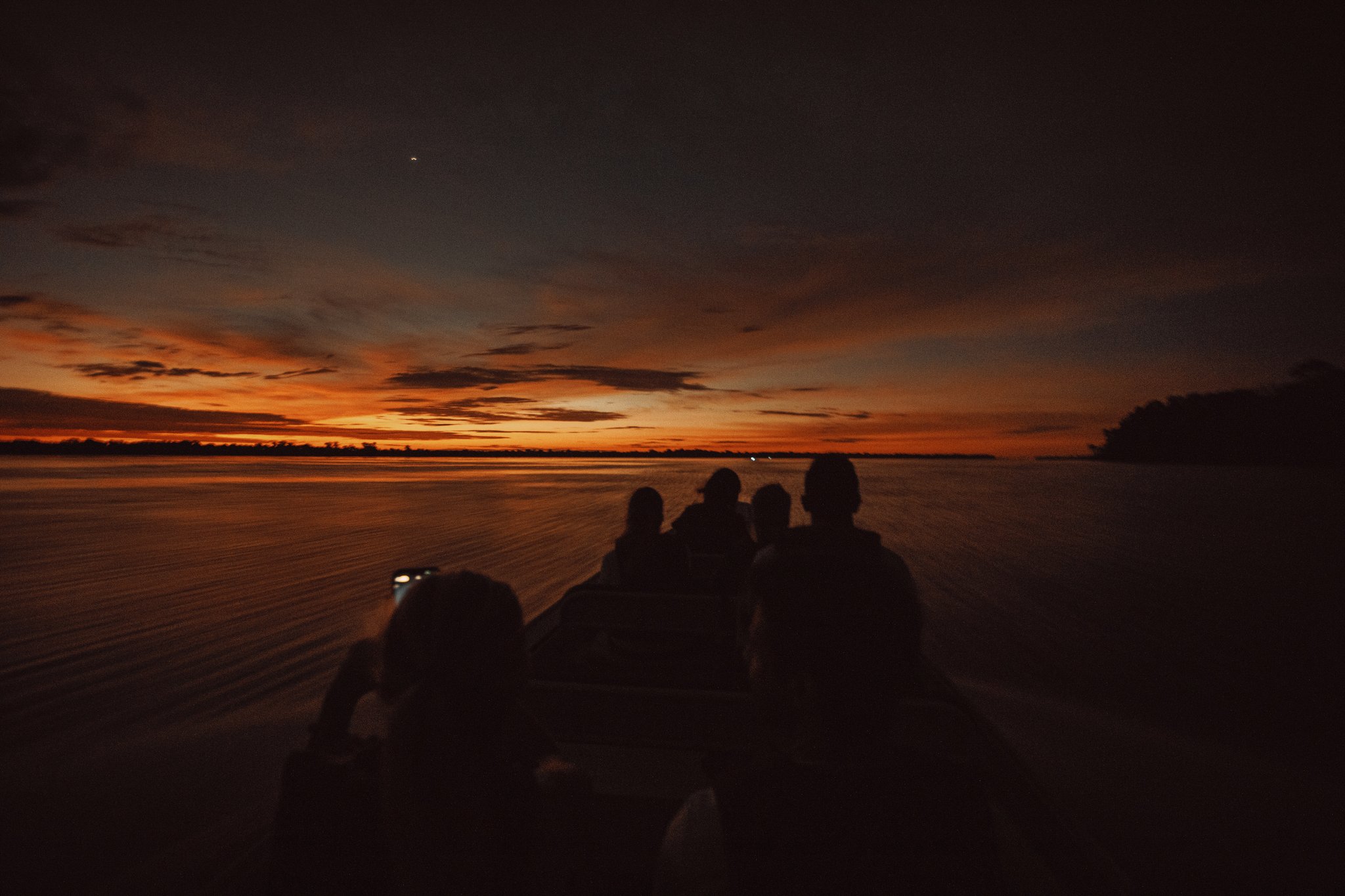 Silhouettes of travellers on a motorboat cutting across a wide Amazon river during a fiery orange-and-red sunset on an amazon rainforest tour in Brazil