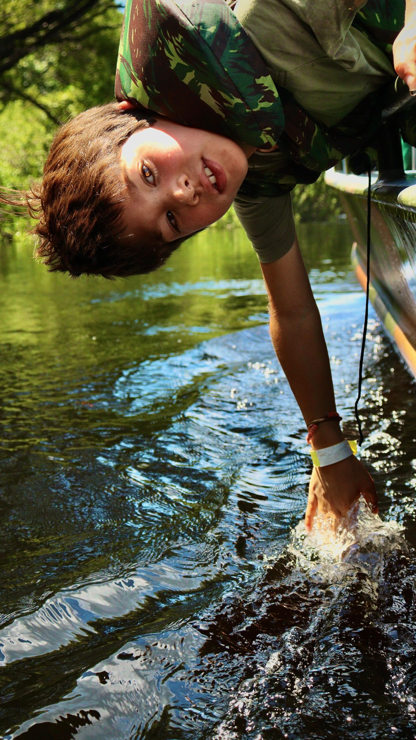 A smiling boy in a camouflage shirt leans over the side of a boat to splash the dark waters of the Amazon River Cruise Brazil route, surrounded by lush green jungle reflections