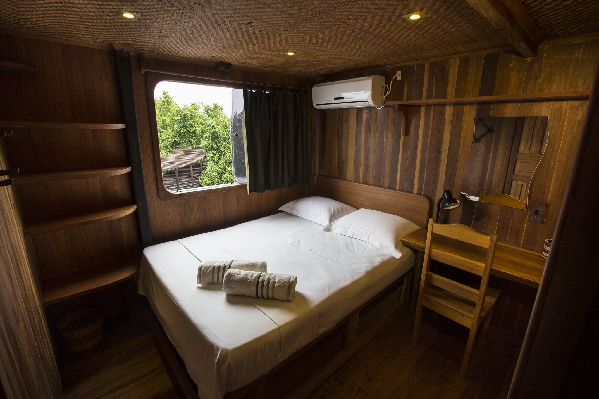 Wood-panelled cabin on an Amazon River Cruise Brazil vessel, featuring a double bed with white linens and rolled towels, open shelving, a desk with lamp, air conditioning unit, and a porthole-style window overlooking green rainforest canopy
