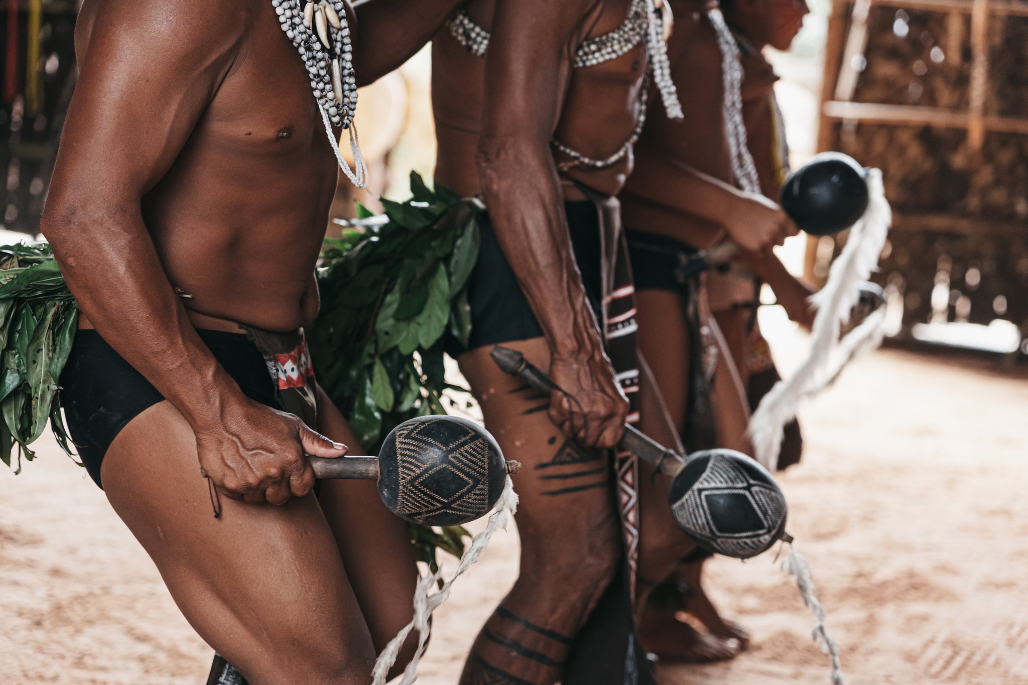 Indigenous men in traditional leaf skirts and bead necklaces holding ornately woven ceremonial rattles during a riverside community visit on an Amazon River Cruise in Brazil