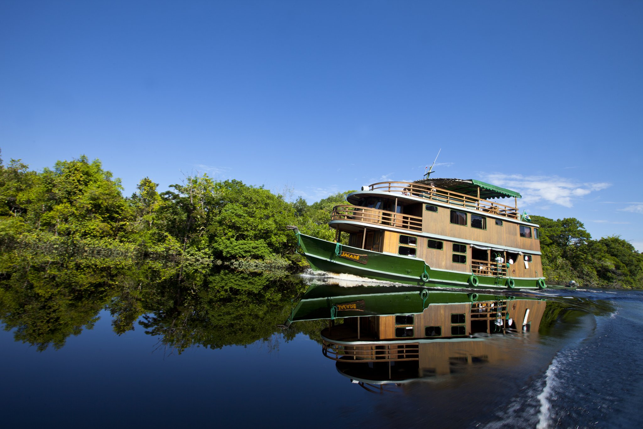 Three decks, open railings, and a river that doubles the sky — the Jacaré sets off into the heart of the Amazon. The Jacaré houseboat cruising a glassy dark-water river on an Amazon River Cruise Brazil, its green-and-timber hull perfectly mirrored in the still water with dense rainforest lining the banks