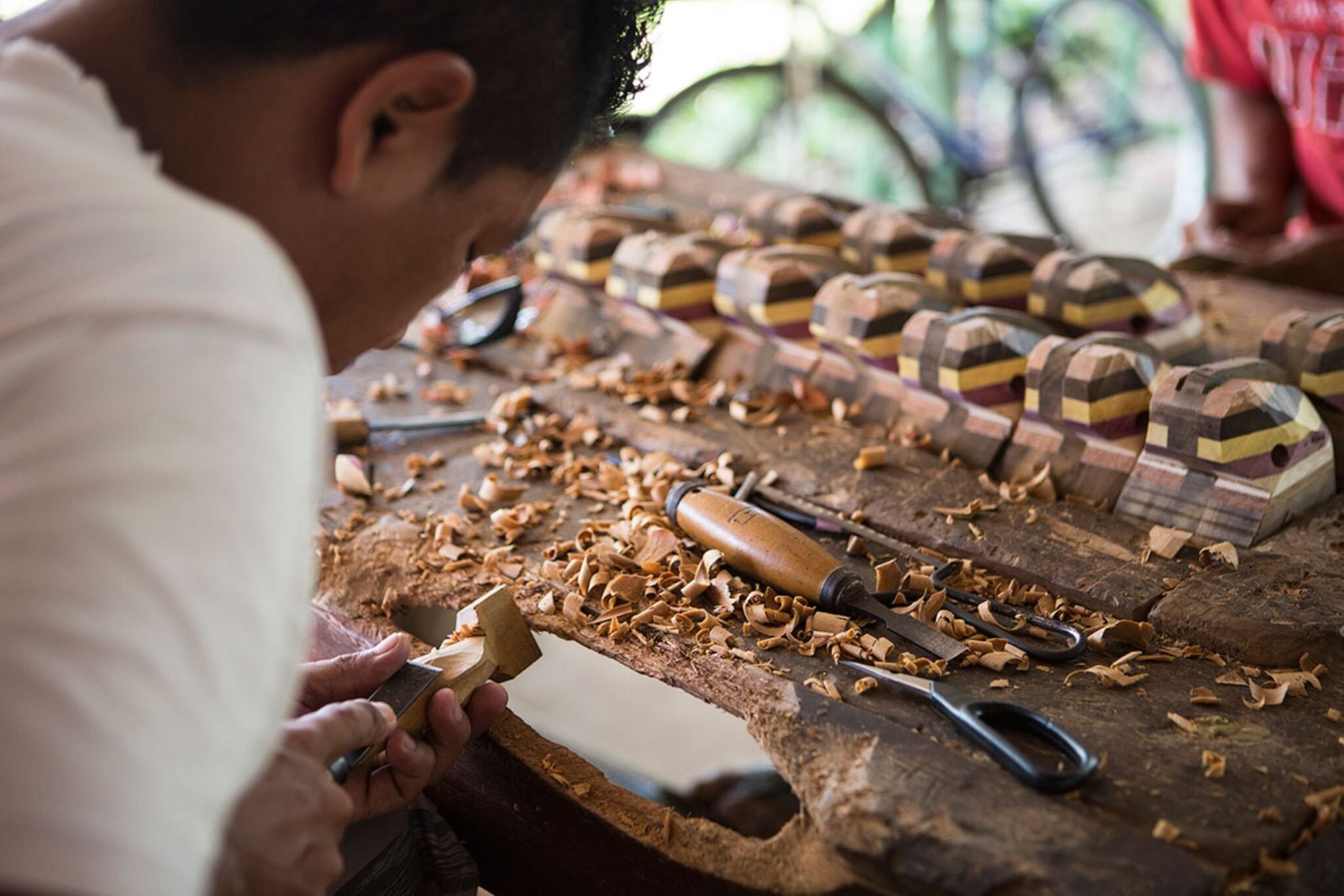Curling shavings, patient hands, and shelves of colour — craft alive in the Amazon. A craftsman carves a small wooden piece by hand on a workbench scattered with fresh shavings, with rows of multicoloured laminated wood figurines lined up behind him — a local art workshop visited on an Amazon River Cruise Brazil itinerary