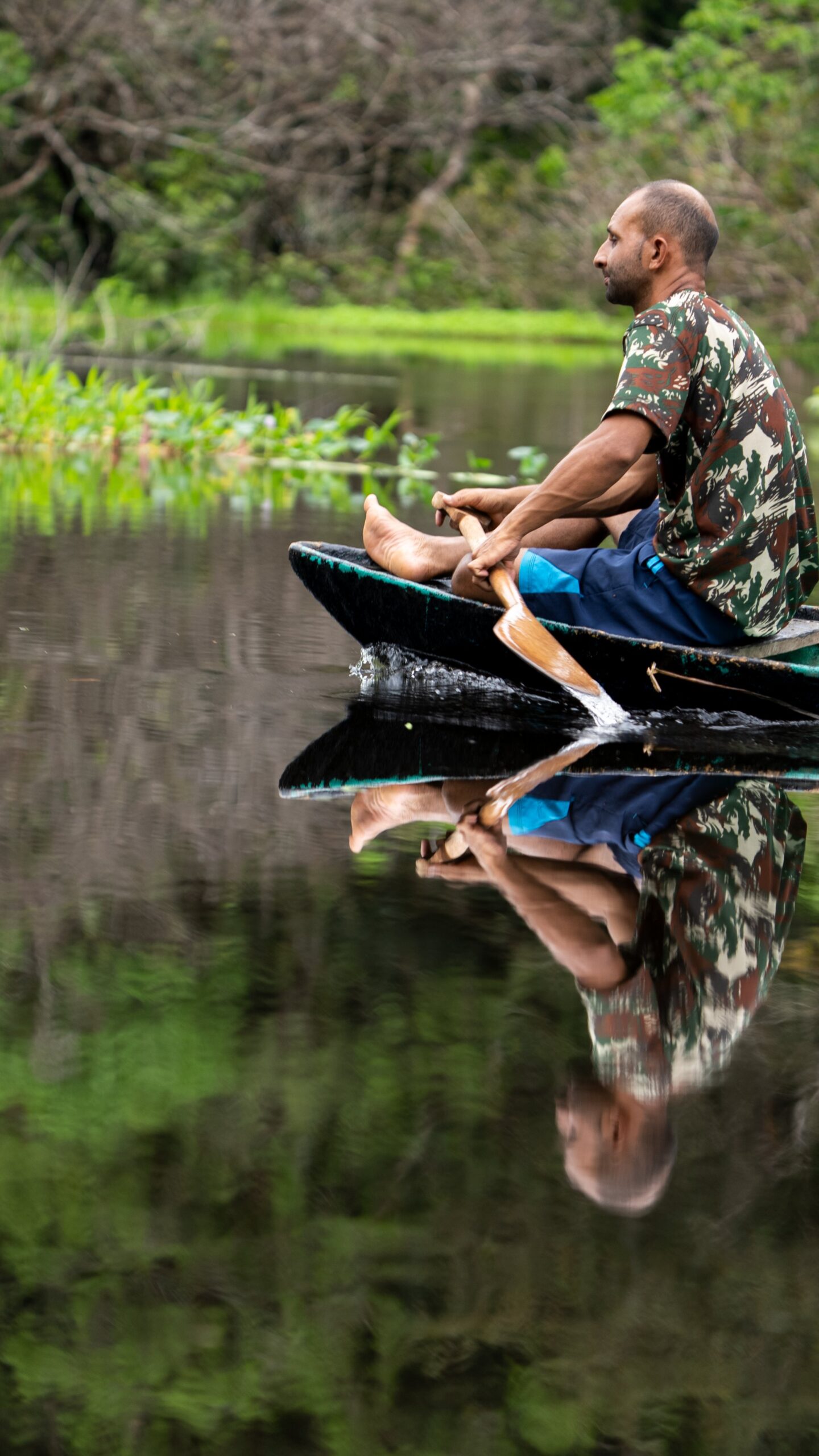 He reads this river the way most of us read a map, effortlessly. A man paddles a traditional wooden canoe across glassy, still water on an Amazon survival tour, his reflection perfectly mirrored on the dark surface beside lush green riverbank vegetation