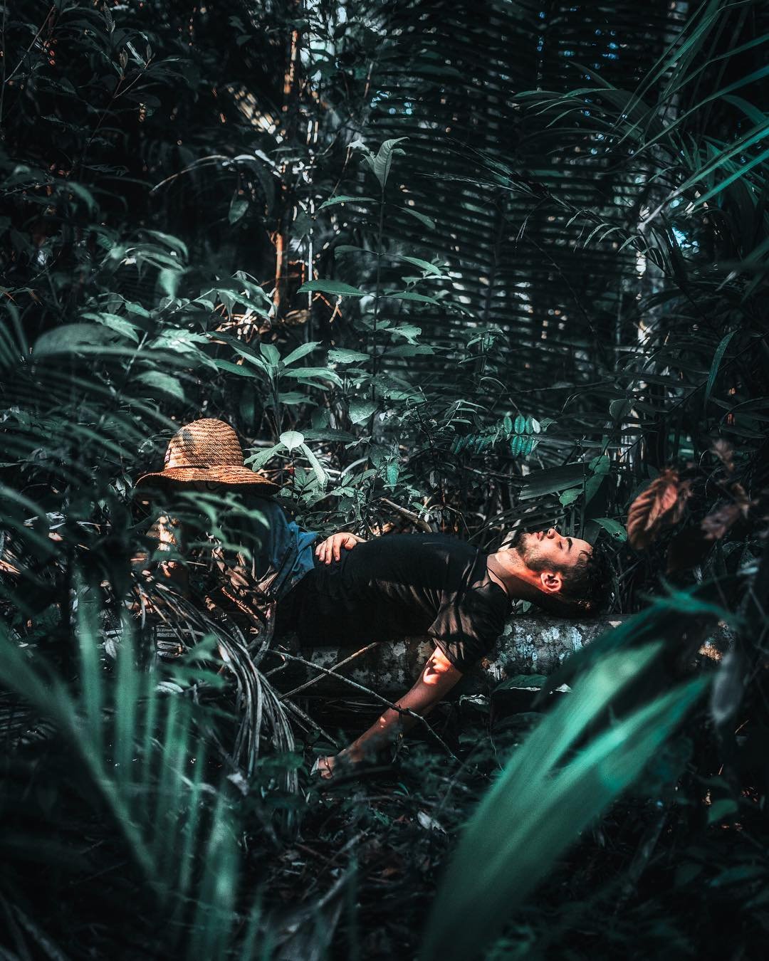 A man reclining on a mossy log deep in dense rainforest undergrowth during an Amazon survival tour, his straw hat resting beside him amid layers of tropical leaves