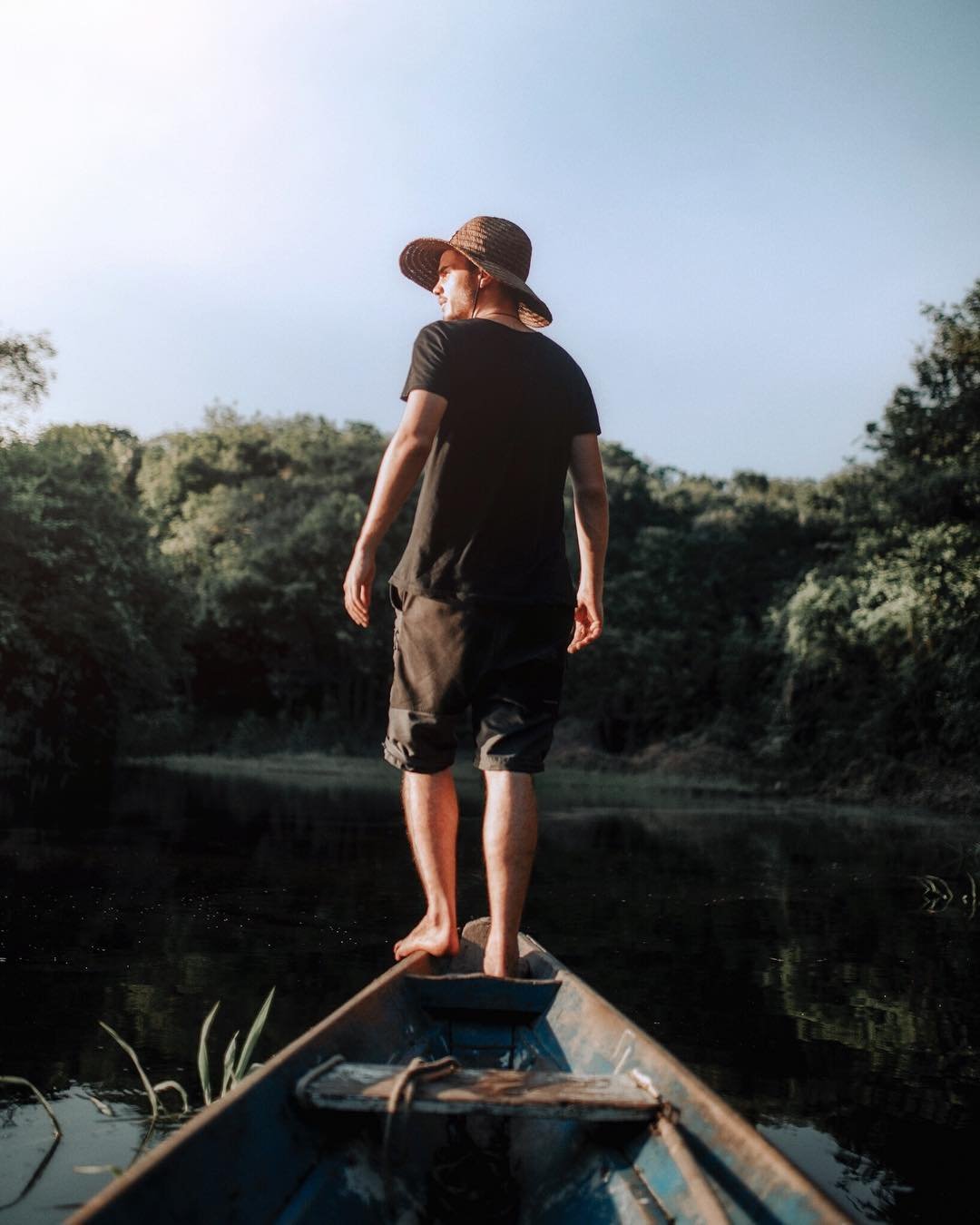 Barefoot on the bow, the river ahead and the forest closing in. A barefoot man in a straw hat stands on the bow of a weathered wooden canoe on an Amazon trek and survival tour, gazing across a calm dark river flanked by dense green forest