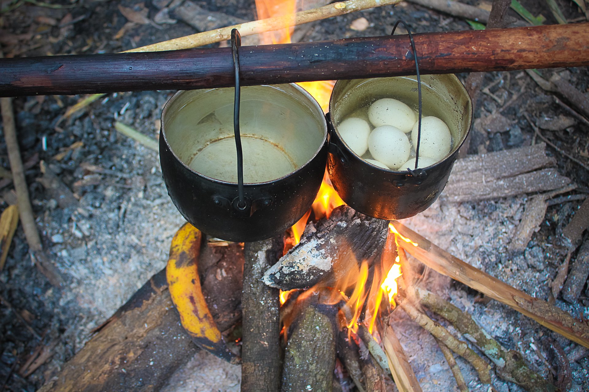 Dinner sorted, no kitchen required. Two blackened pots hanging over an open wood fire on a jungle survival Brazil camp, one holding eggs boiling in water