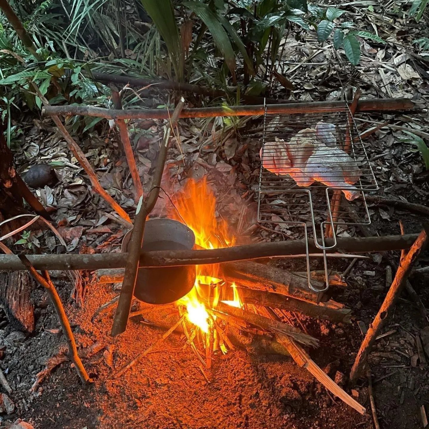A clay pot and wire grill balanced over an open wood fire on a rustic bamboo frame deep in the jungle, part of an Amazon trek and survival tour camp meal