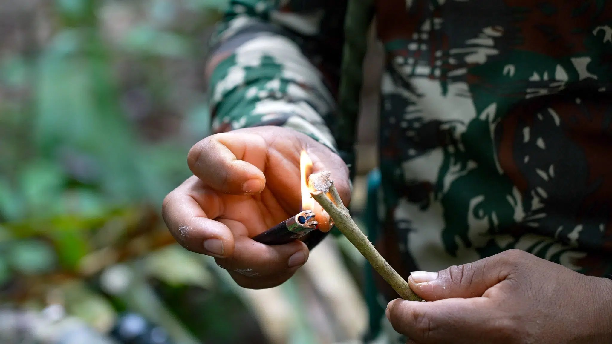 Old knowledge, live flame, green forest all around. A guide in camouflage clothing lights a green stick using a lighter during an Amazon trek and survival tour, demonstrating a bushcraft fire-starting technique in the rainforest