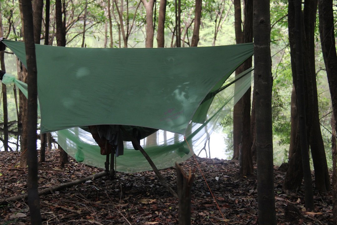 A hammock wrapped in a white mosquito net hangs beneath a rustic thatched palm shelter in dense rainforest, part of an Amazon trek and survival tour wild camp