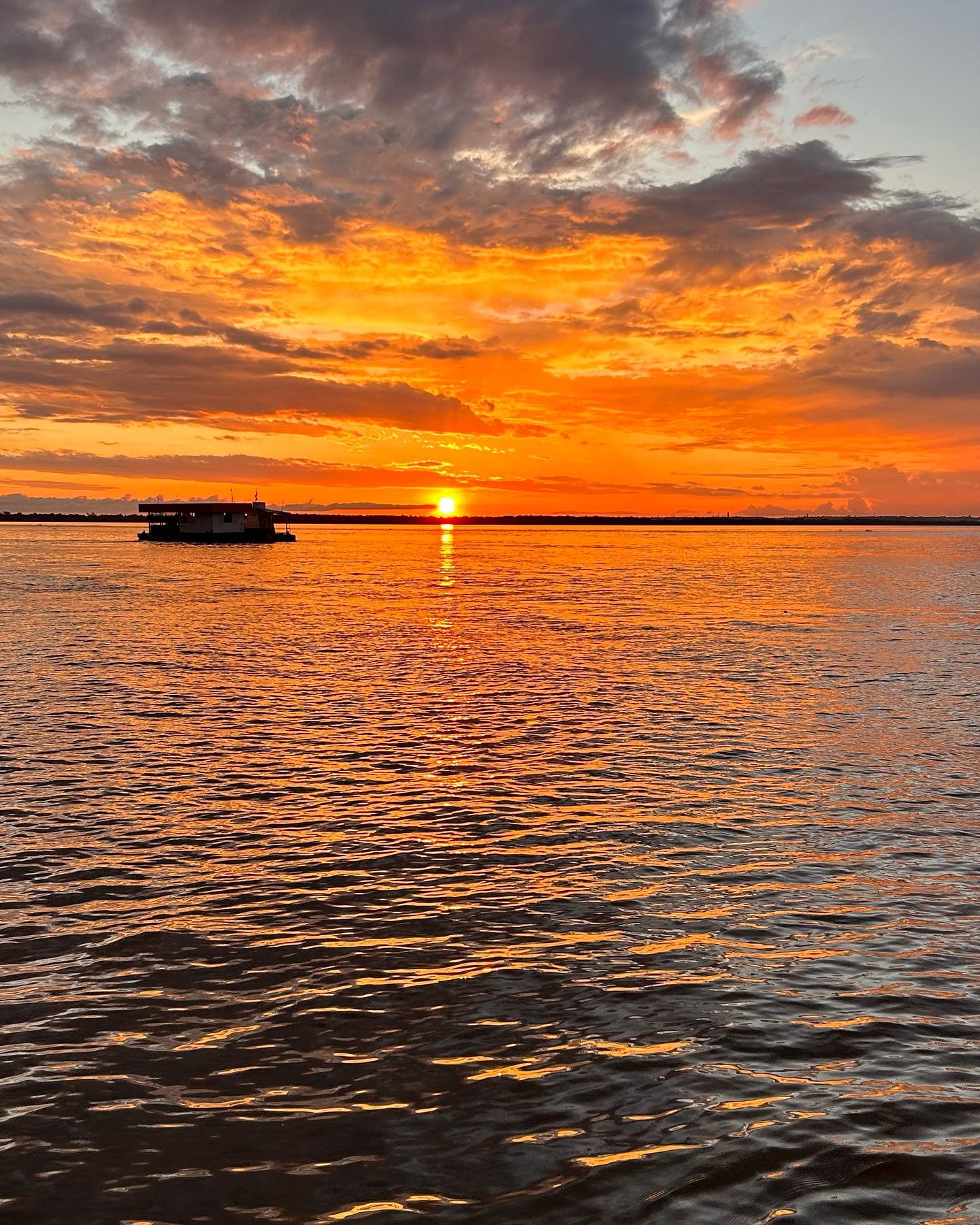 A houseboat silhouetted against a blazing orange and red sunset reflected across a wide Amazonian river, the kind of scene encountered on an Amazon trek and survival tour departing from Manaus