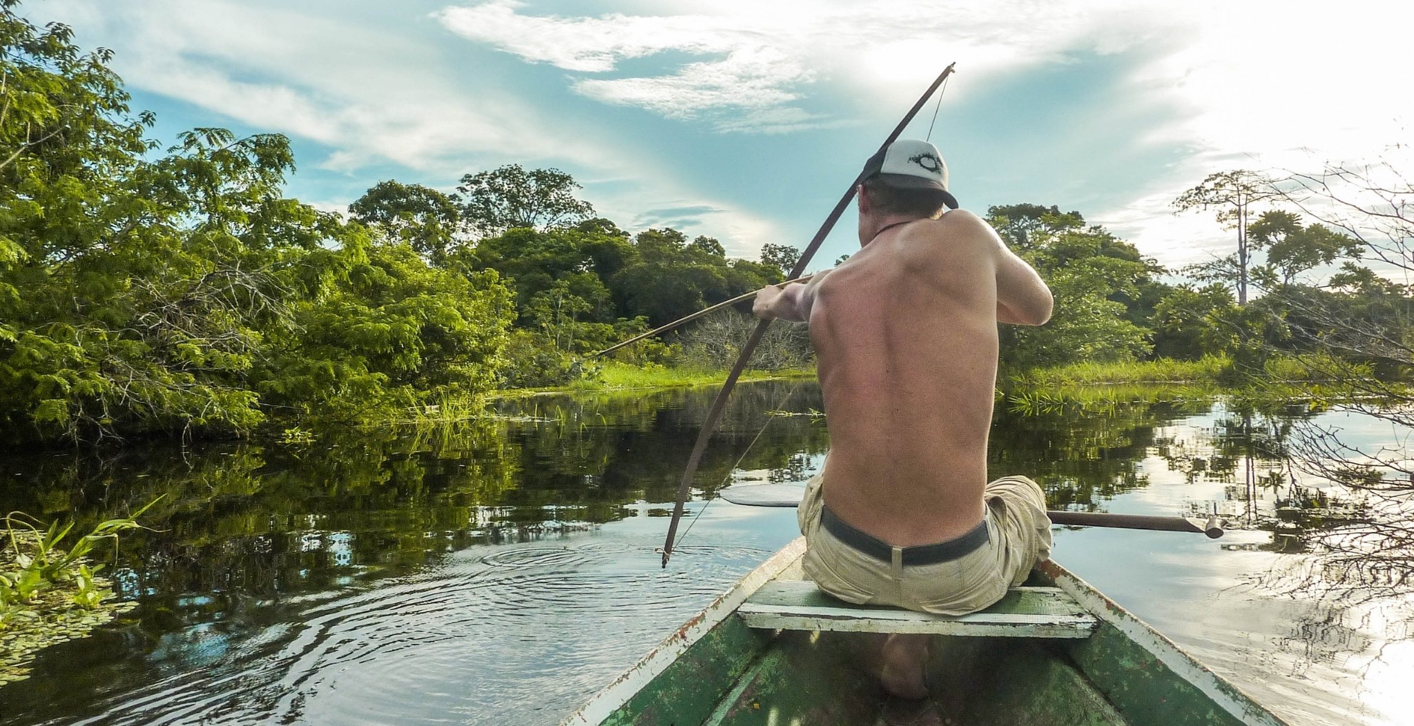 A shirtless man kneels at the bow of a weathered wooden canoe, drawing back a long spear to fish on a calm jungle waterway lined with dense green tropical forest — a raw moment from an Amazon trek and survival tour