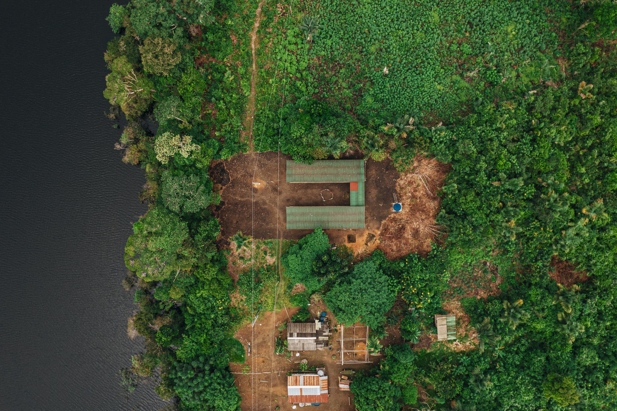 Aerial drone view of Anavilhanas Jungle Lodge Brazil, with green-roofed buildings nestled in dense rainforest canopy beside a dark Rio Negro waterway and a small riverside community below