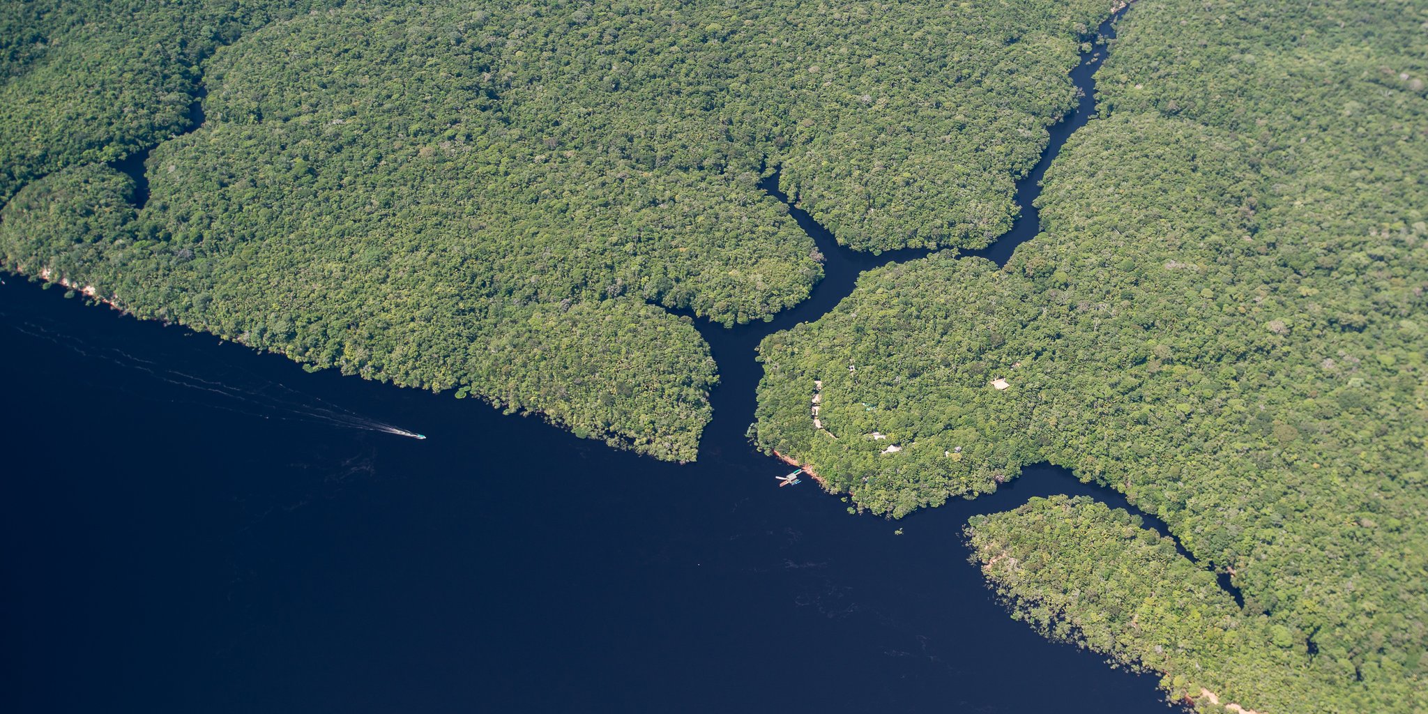 Aerial view of Anavilhanas Jungle Lodge Brazil nestled at the edge of dark Rio Negro waters, surrounded by an unbroken carpet of dense Amazon rainforest canopy with narrow channels threading between forested islands, a small boat leaving a wake on the river below