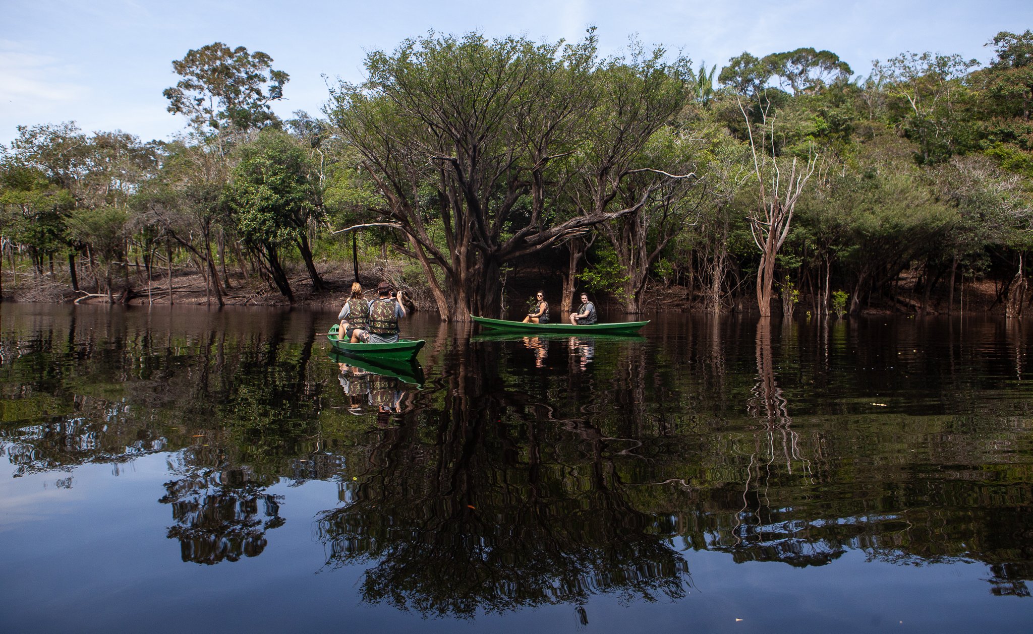 Three travellers in a green canoe gliding through a flooded forest on dark mirror-still water at Anavilhanas Jungle Lodge Brazil, surrounded by partially submerged trees and their perfect reflections