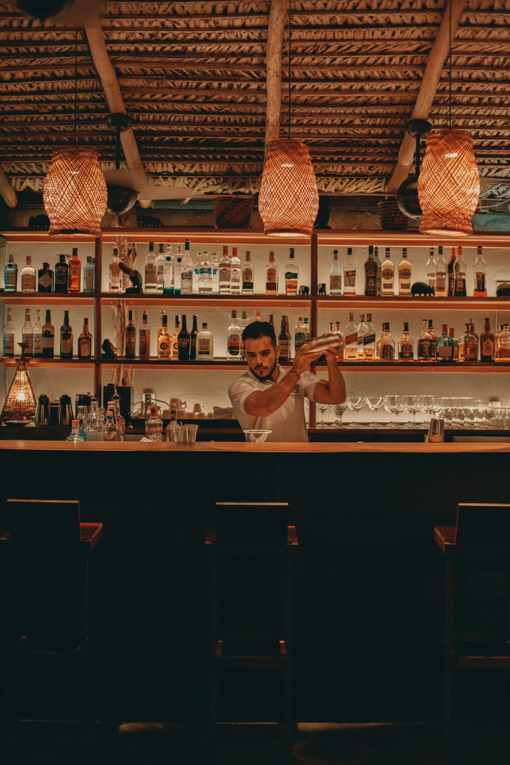Skilled hands, glowing rattan, and a well-stocked bar — the night is just getting started. A bartender shaking a cocktail shaker behind the bar at Anavilhanas Jungle Lodge Brazil, beneath warm woven rattan pendant lights and shelves lined with spirits