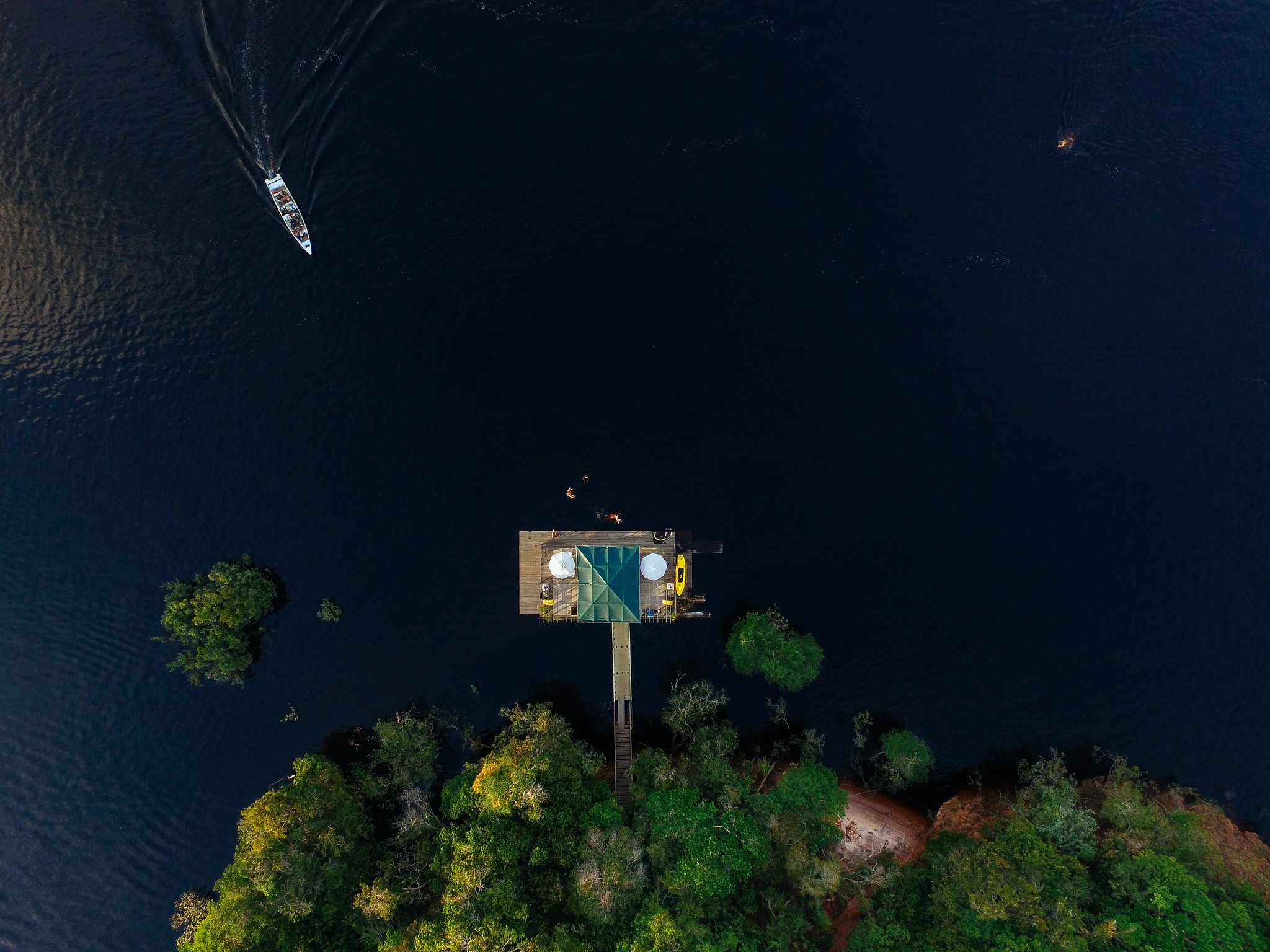 Aerial drone view of the Anavilhanas Jungle Lodge Brazil floating bar and deck extending over the dark waters of the Rio Negro, with a speedboat cutting across the river and dense rainforest canopy visible at the bottom of the frame