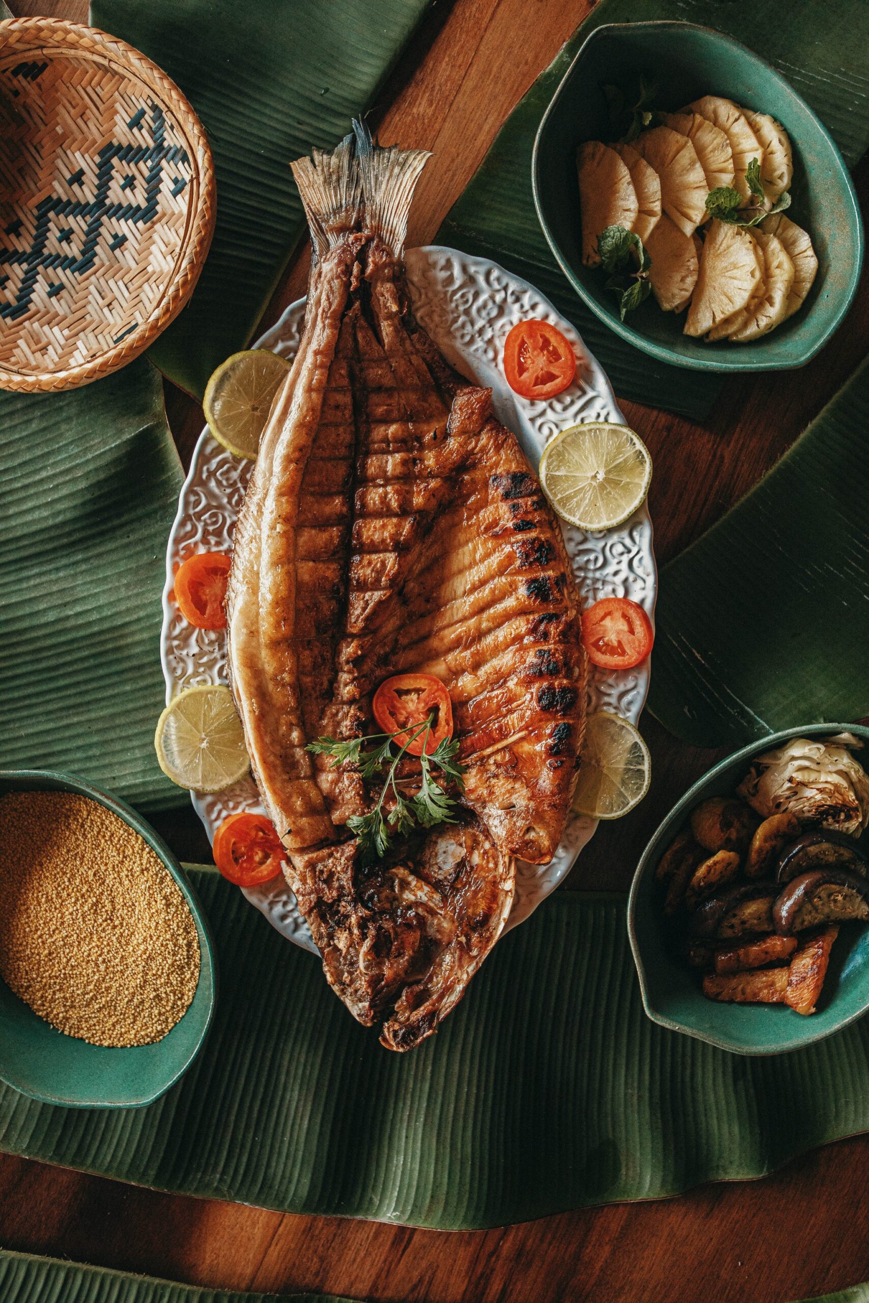 A whole grilled fish served on a decorative white platter with lime and tomato slices at Anavilhanas Jungle Lodge Brazil, surrounded by side bowls of pineapple, roasted mushrooms and golden seeds on banana leaves