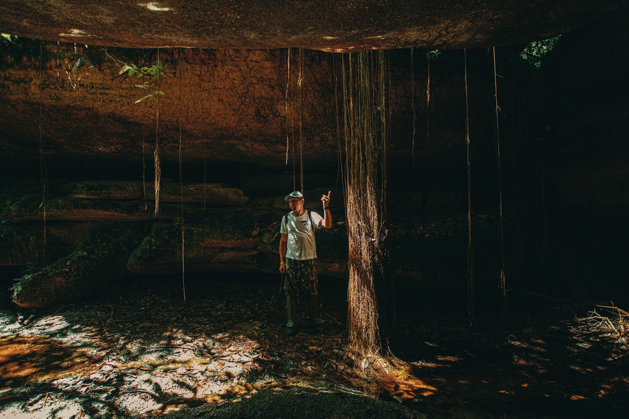 A guide points upward beneath a massive ancient rock overhang at the Madadá Caves, Anavilhanas Jungle Lodge Brazil, with long aerial roots cascading from the ceiling in dramatic golden light