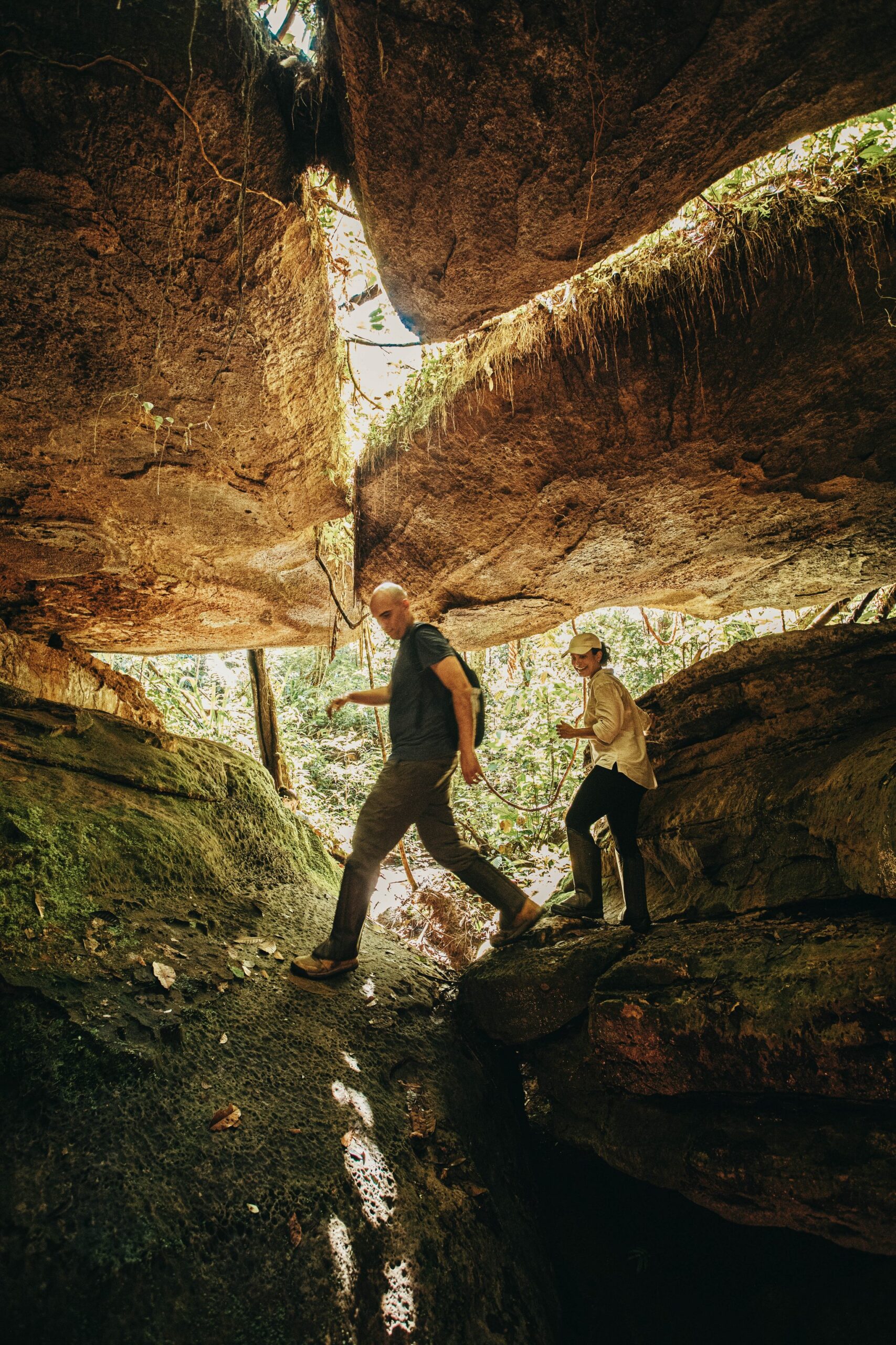 Two hikers duck beneath a massive sandstone overhang at the Madadá Caves during an Anavilhanas Jungle Lodge Brazil excursion, sunlit jungle visible through the rock opening behind them