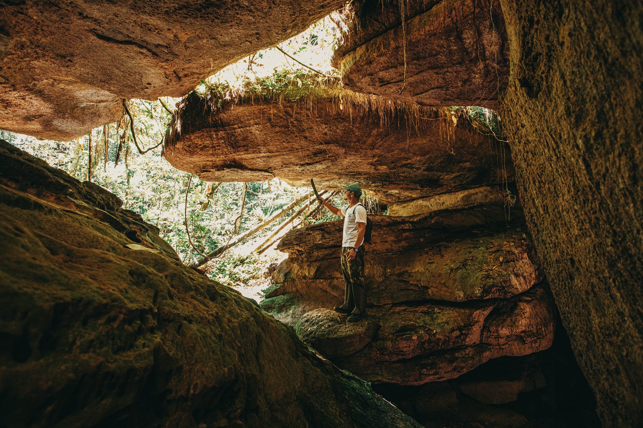 Seven hundred million years in the making — a guide reads the rock. A guide stands inside the ancient sandstone cave formations of the Madadá Caves on a hiking excursion from Anavilhanas Jungle Lodge Brazil, holding a stick and gazing up at the dramatic overhanging rock ceiling draped with hanging roots