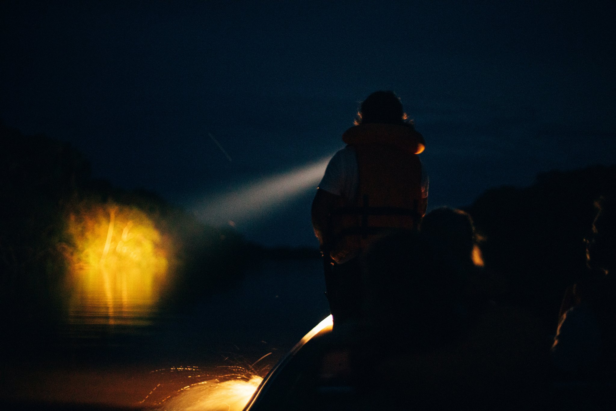 A silhouetted guide in a life vest sweeps a spotlight across dark river water during a night tour at Anavilhanas Jungle Lodge Brazil, with a warm yellow lamp glowing at the bow