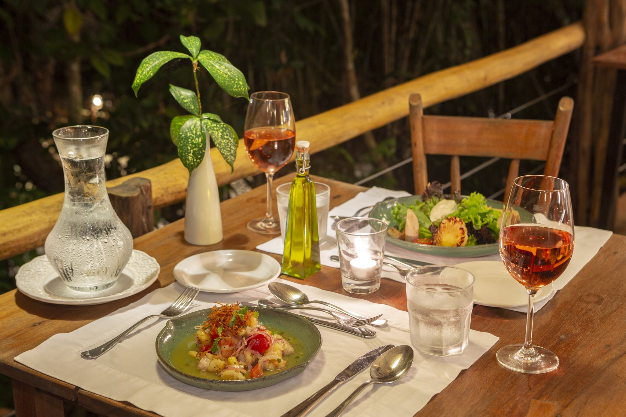 A candlelit outdoor dining table at Anavilhanas Jungle Lodge Brazil set with rosé wine, a green ceramic plate of seafood starter, fresh salad, olive oil and a tropical leaf centrepiece, overlooking the jungle on a bamboo-railed terrace