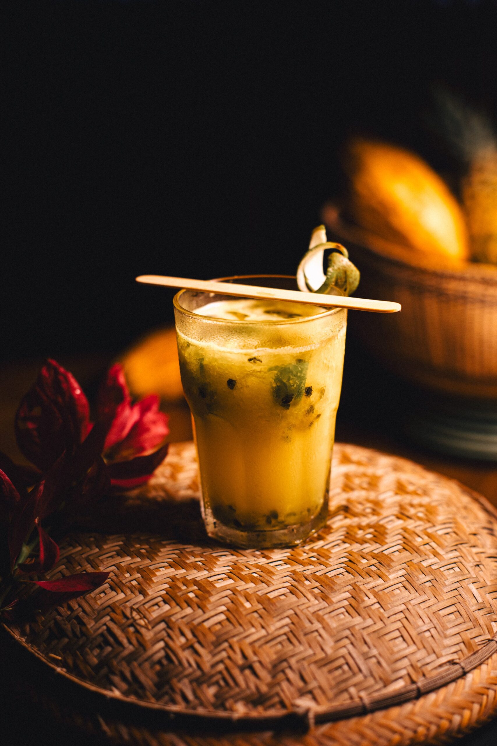 A golden passion fruit cocktail with visible seeds served in a glass on a woven rattan tray at Anavilhanas Jungle Lodge Brazil, garnished with a wooden stirrer and a curled tropical leaf, with a basket of yellow fruit softly lit in the background