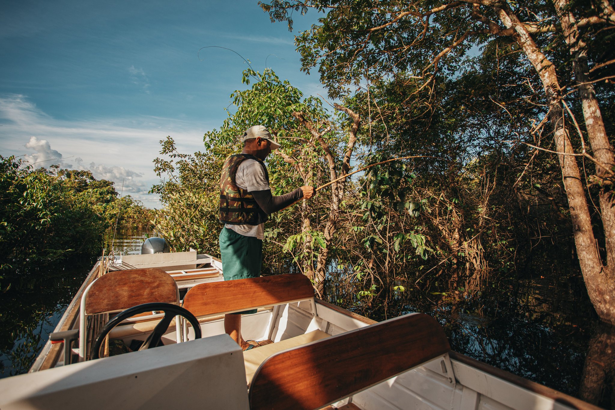 A guide stands at the bow of a small motorboat, fishing rod in hand, casting toward dense riverside vegetation during a recreational fishing excursion at Anavilhanas Jungle Lodge Brazil