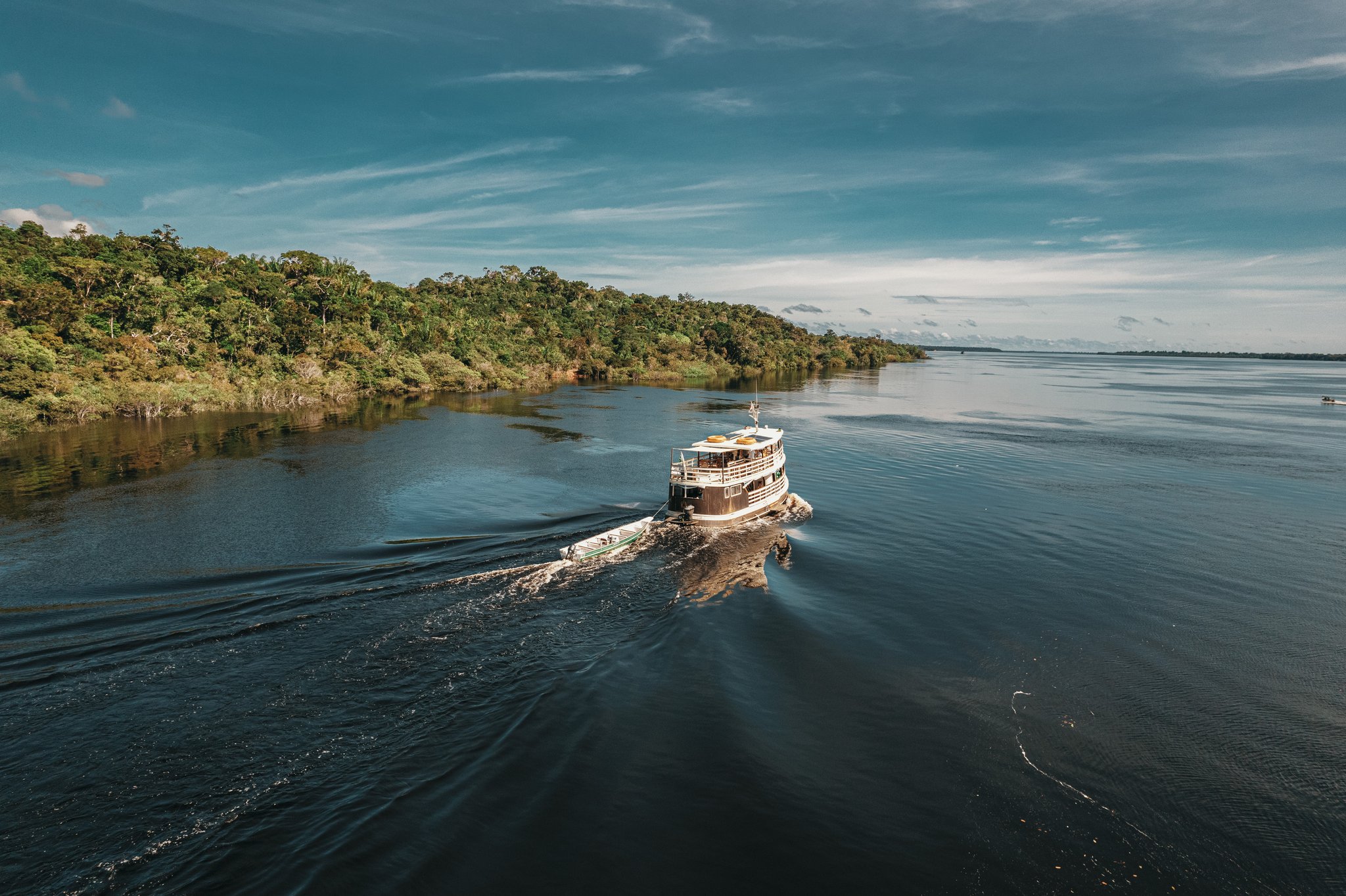 Open water, endless forest — the Rio Negro stretches out ahead. A two-deck wooden expedition boat cruises the dark waters of the Rio Negro past dense Amazon rainforest on a full-day Anavilhanas Jungle Lodge Brazil excursion