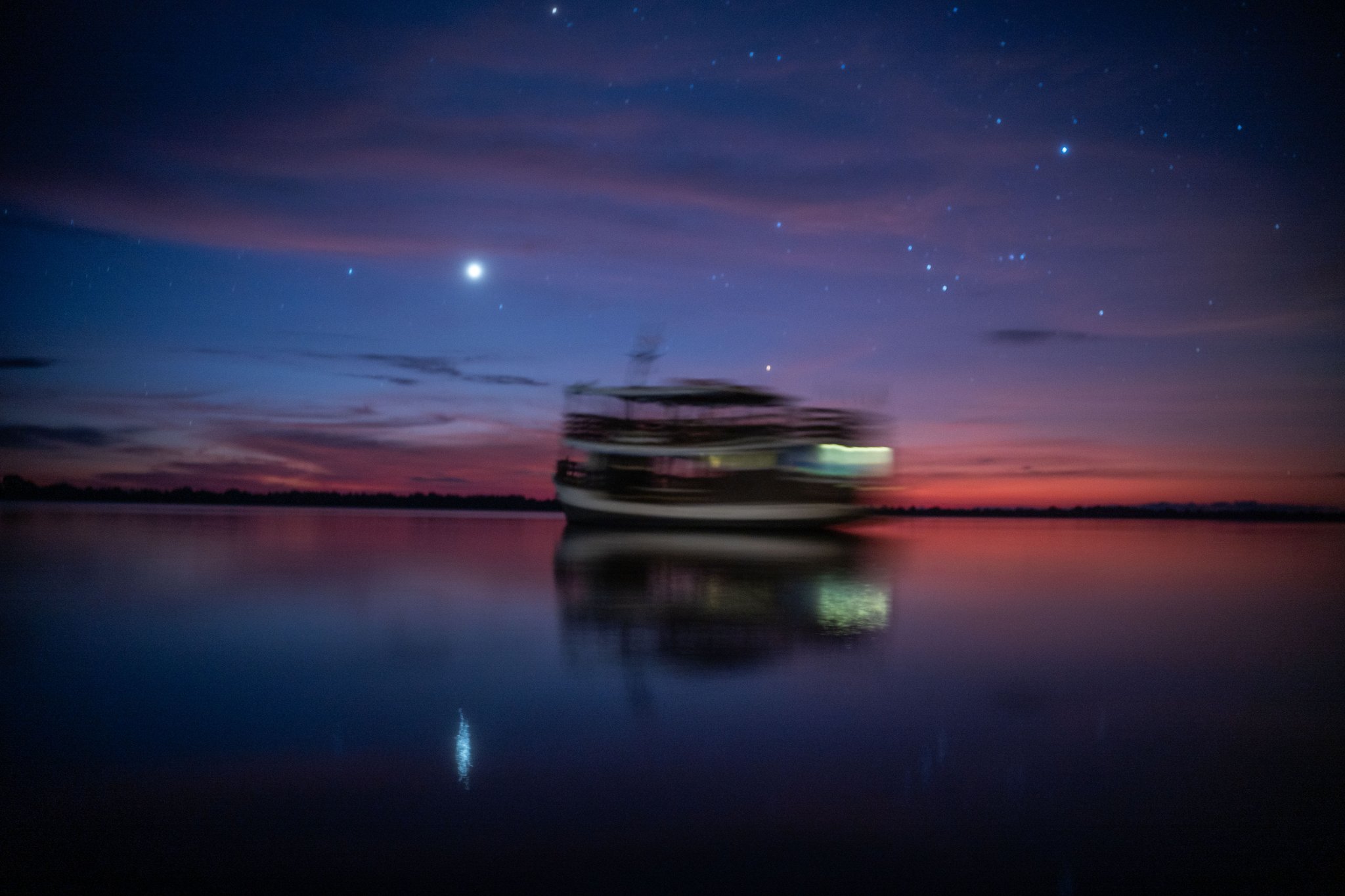 A multi-deck river vessel gliding across mirror-still dark water at twilight under a deep violet and crimson sky at Anavilhanas Jungle Lodge Brazil, with stars emerging above and the boat's lights reflected below