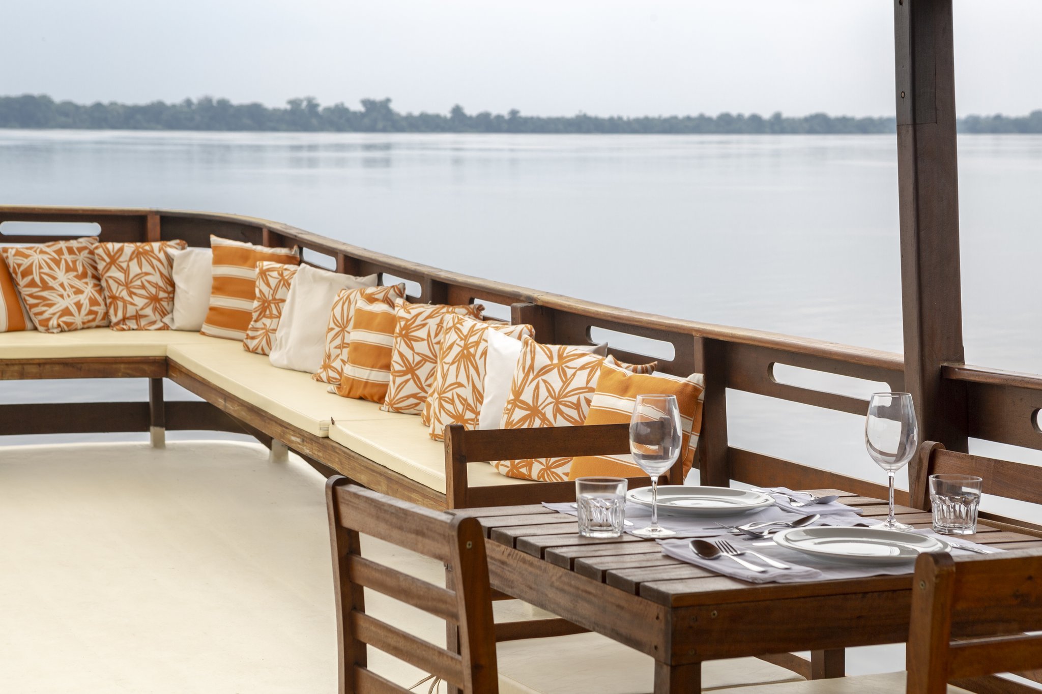 A set dining table with wine glasses and cutlery on the open deck of a boat at Anavilhanas Jungle Lodge Brazil, with a cushioned lounge bench and a wide river stretching to the forested horizon