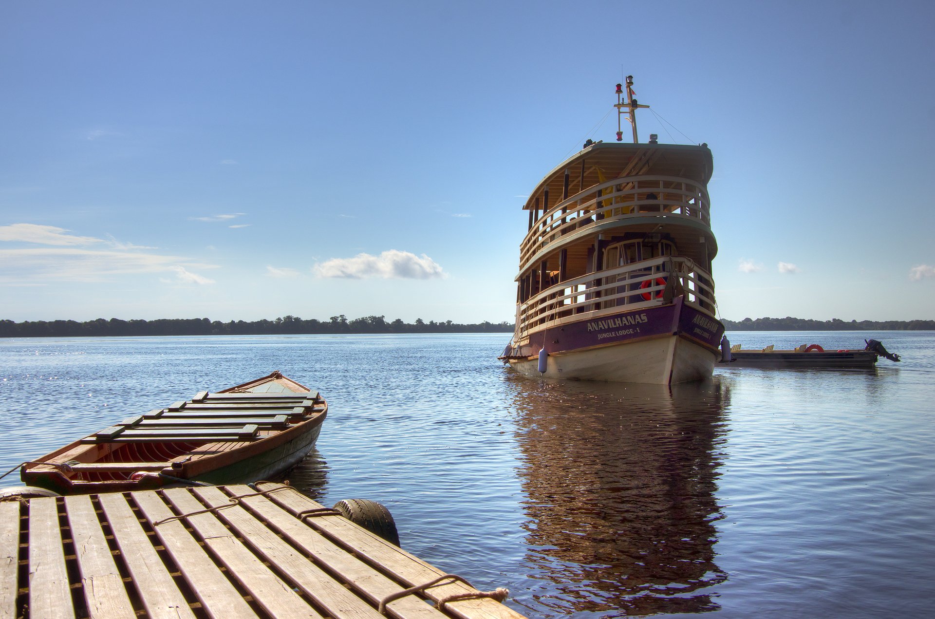 Your floating base camp, ready and waiting on the Rio Negro. The Anavilhanas Jungle Lodge Brazil riverboat moored on the calm black waters of the Rio Negro, viewed from a wooden dock with a traditional wooden canoe in the foreground