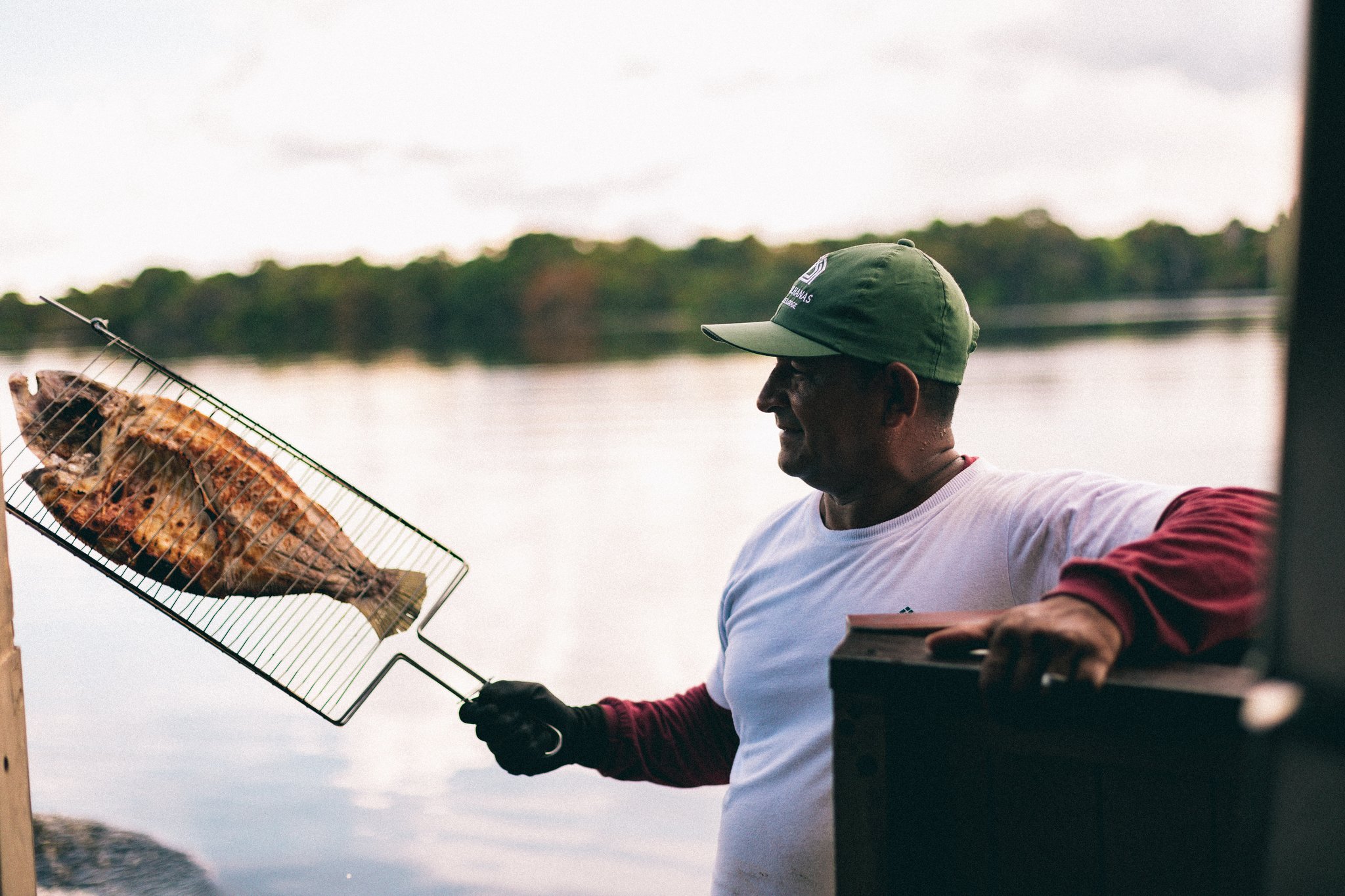Fresh from the river, straight to the grill — lunch doesn't get more Amazon than this. A smiling man in a green cap holds a whole grilled fish in a wire basket over the Rio Negro at Anavilhanas Jungle Lodge Brazil, dense rainforest reflected in the calm water behind him