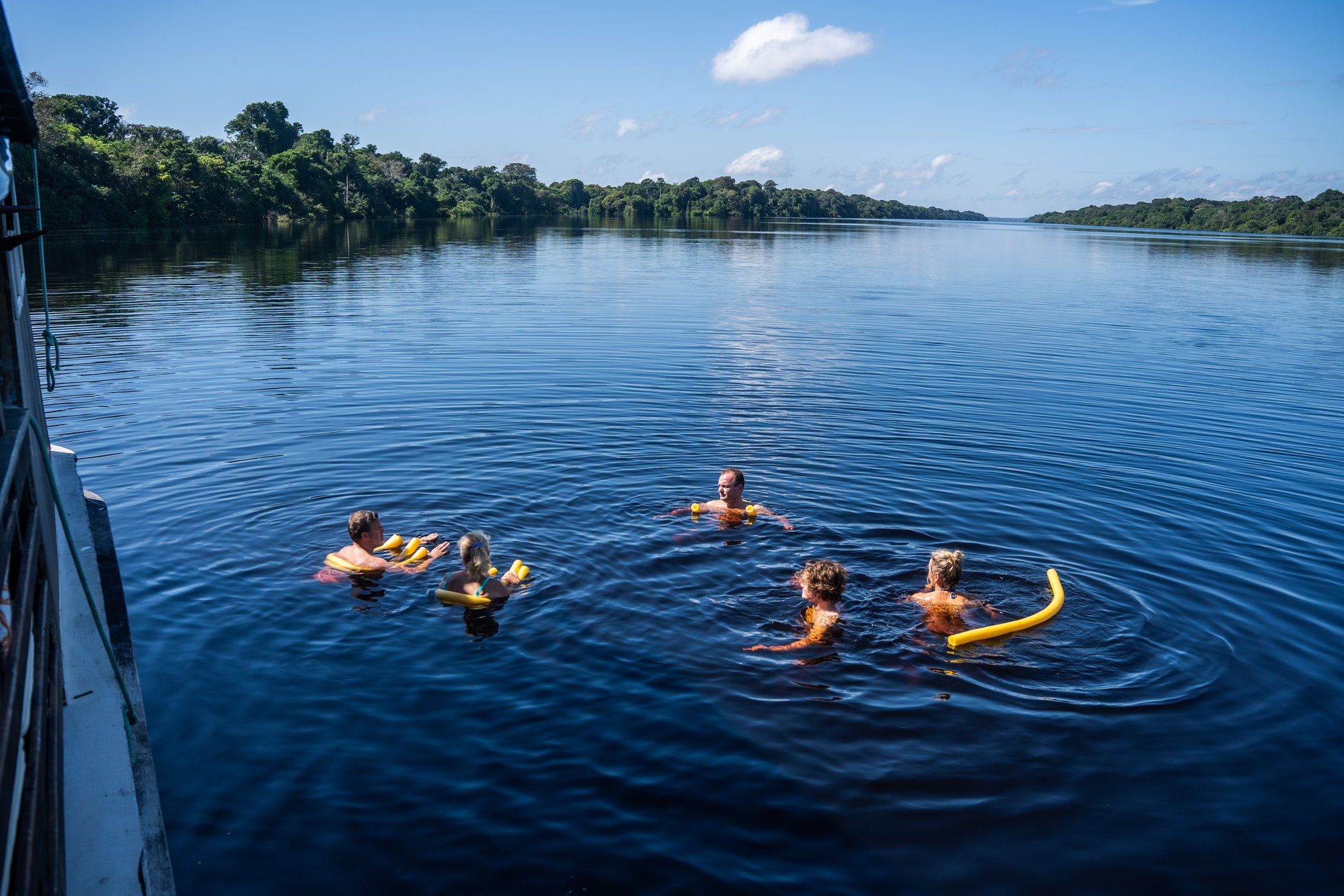 Five guests swimming in the dark waters of the Rio Negro beside a traditional boat at Anavilhanas Jungle Lodge Brazil, with yellow foam noodles and dense rainforest lining both riverbanks under a blue sky