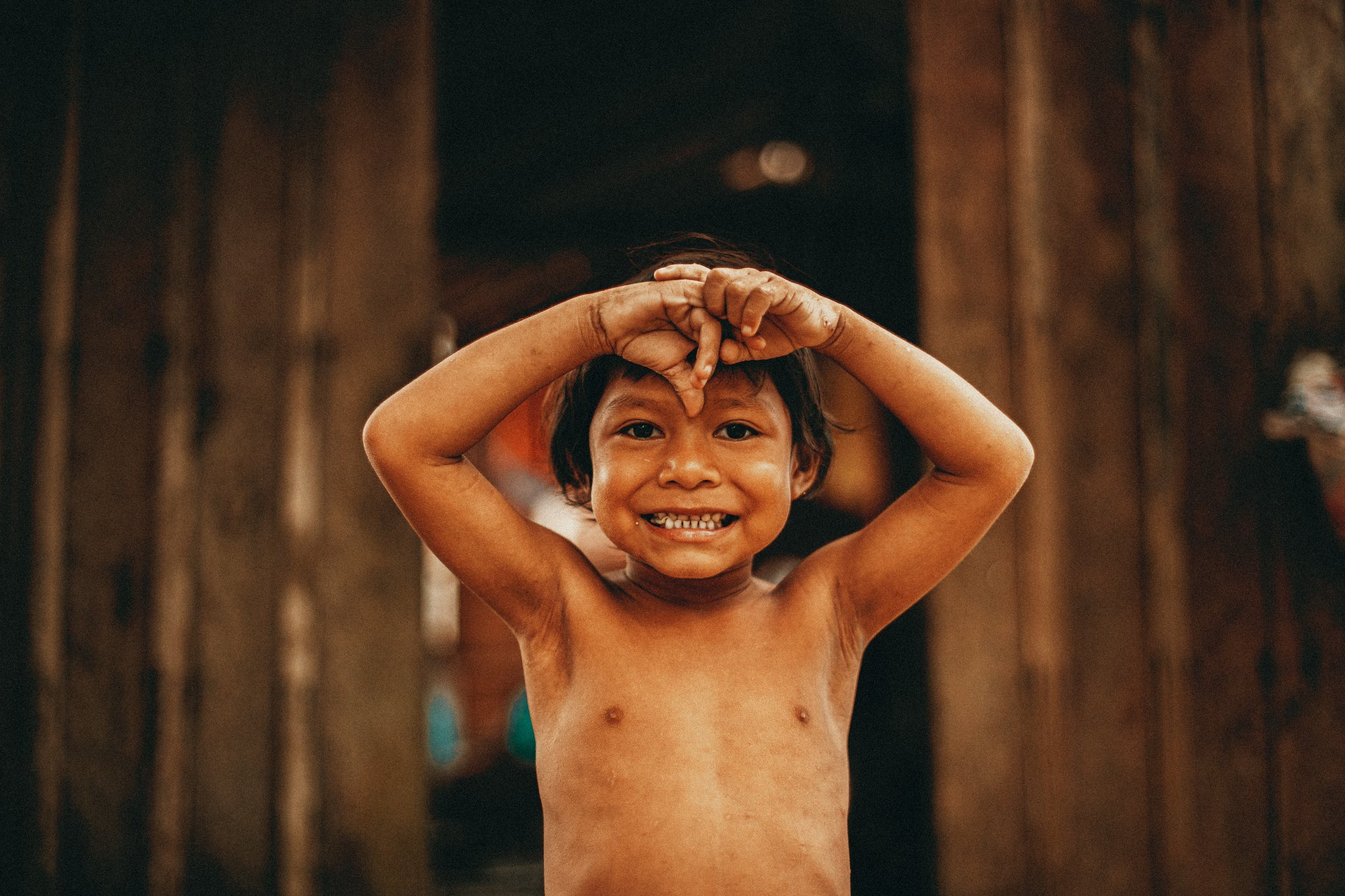 A laughing child in a riverside community near Anavilhanas Jungle Lodge Brazil, hands raised playfully above their head in front of a wooden wall