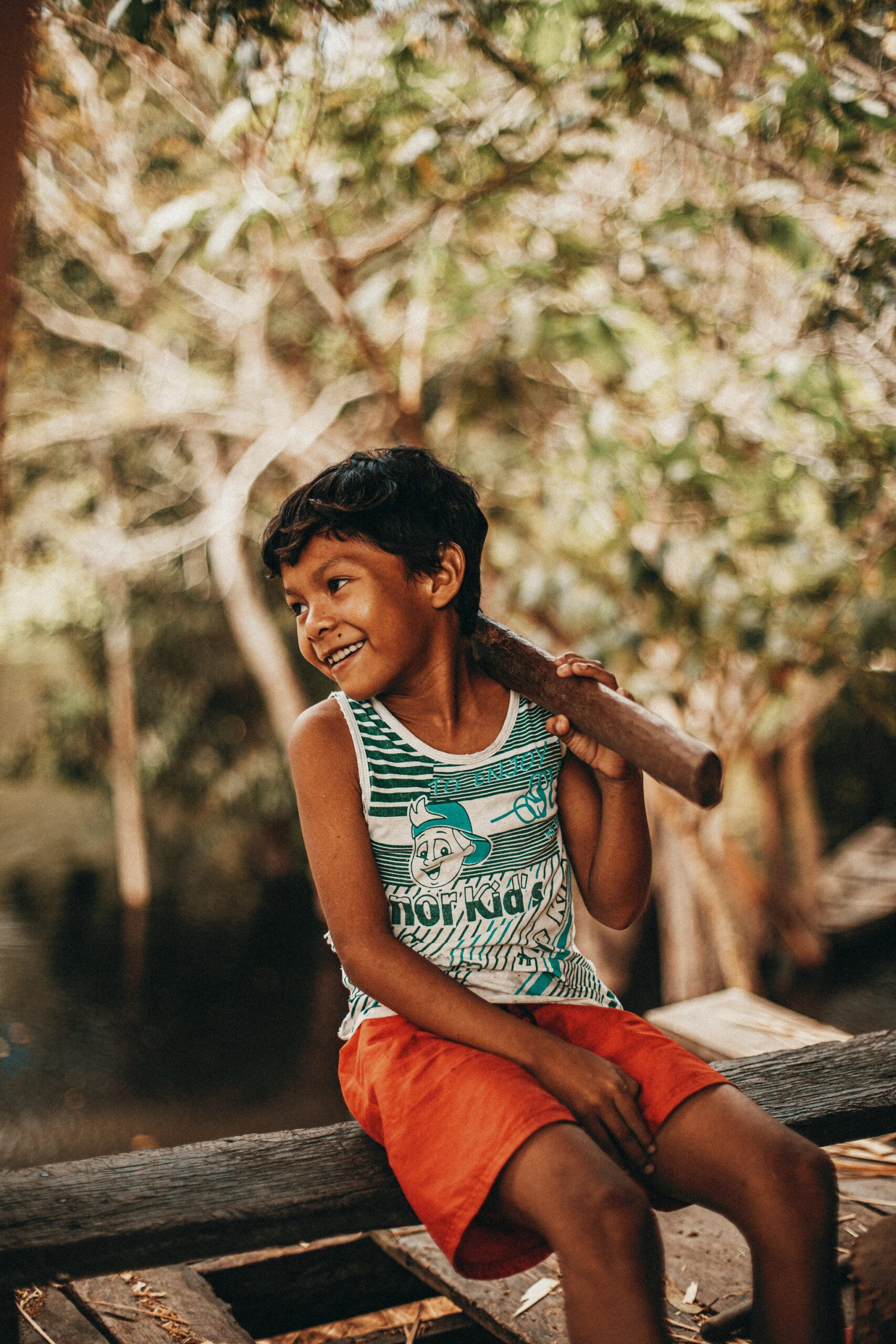 Pure joy, caught mid-grin in a riverside village. A smiling boy sits on a wooden ledge holding a stick over his shoulder during a riverside community visit at Anavilhanas Jungle Lodge Brazil, with sun-dappled trees behind him