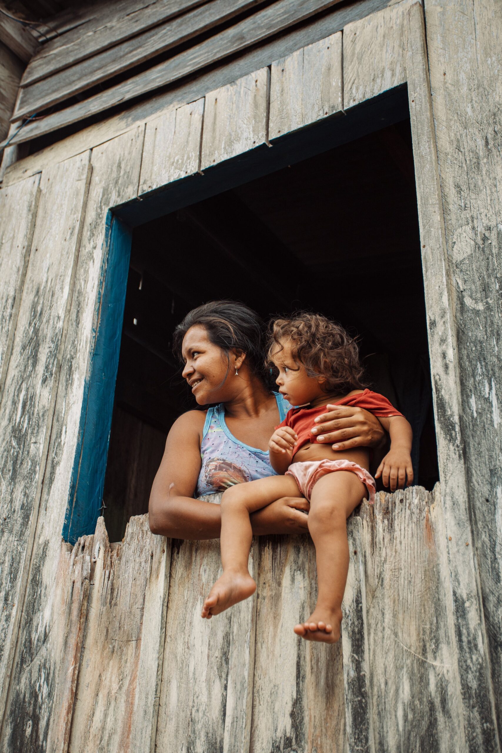 A smiling woman holds a barefoot toddler on the sill of a weathered wooden window during a riverside community visit near Anavilhanas Jungle Lodge Brazil