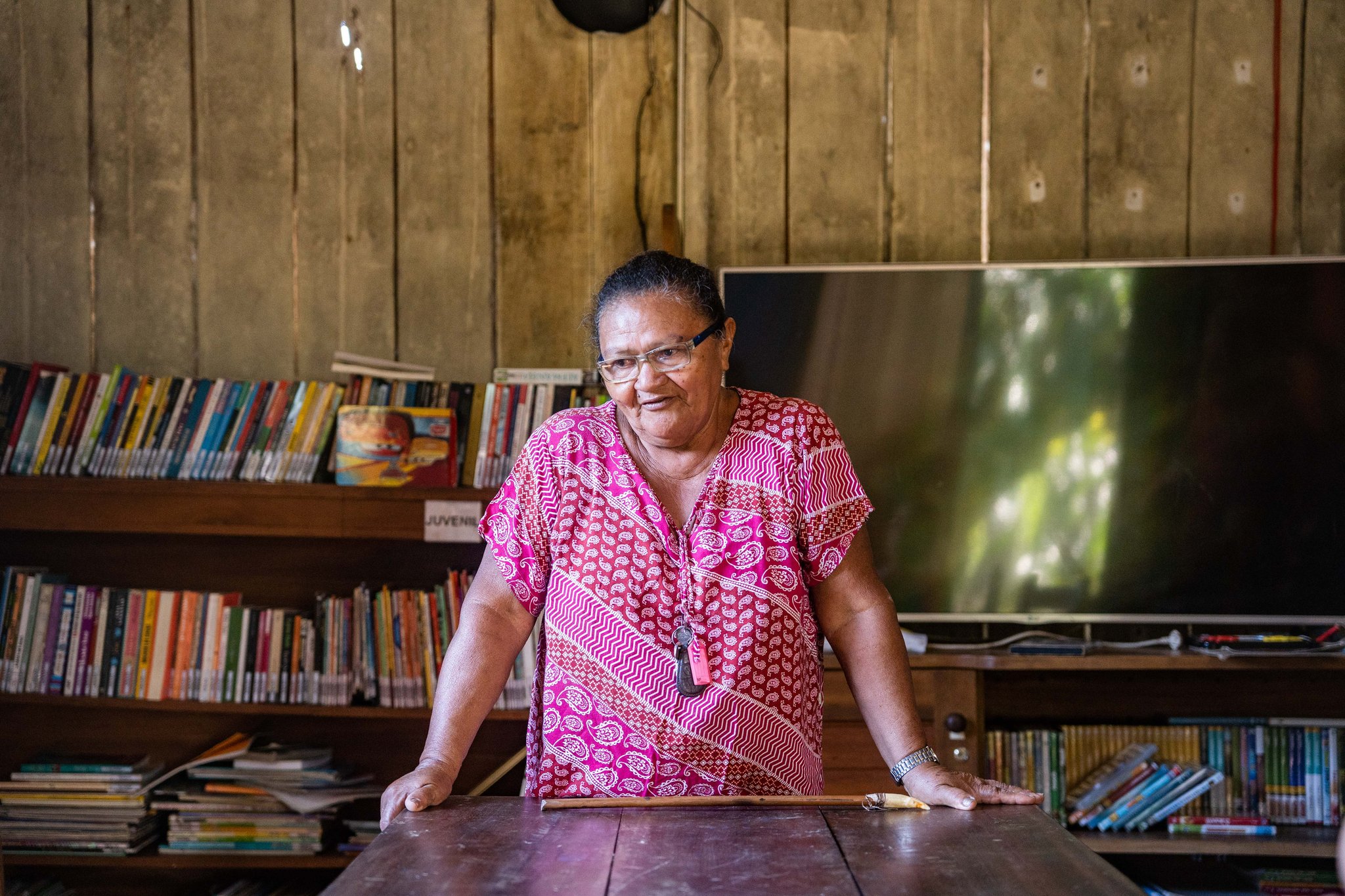 A local woman in a pink patterned dress stands at a wooden table inside a community library near Anavilhanas Jungle Lodge Brazil, with colourful bookshelves lining the timber walls behind her