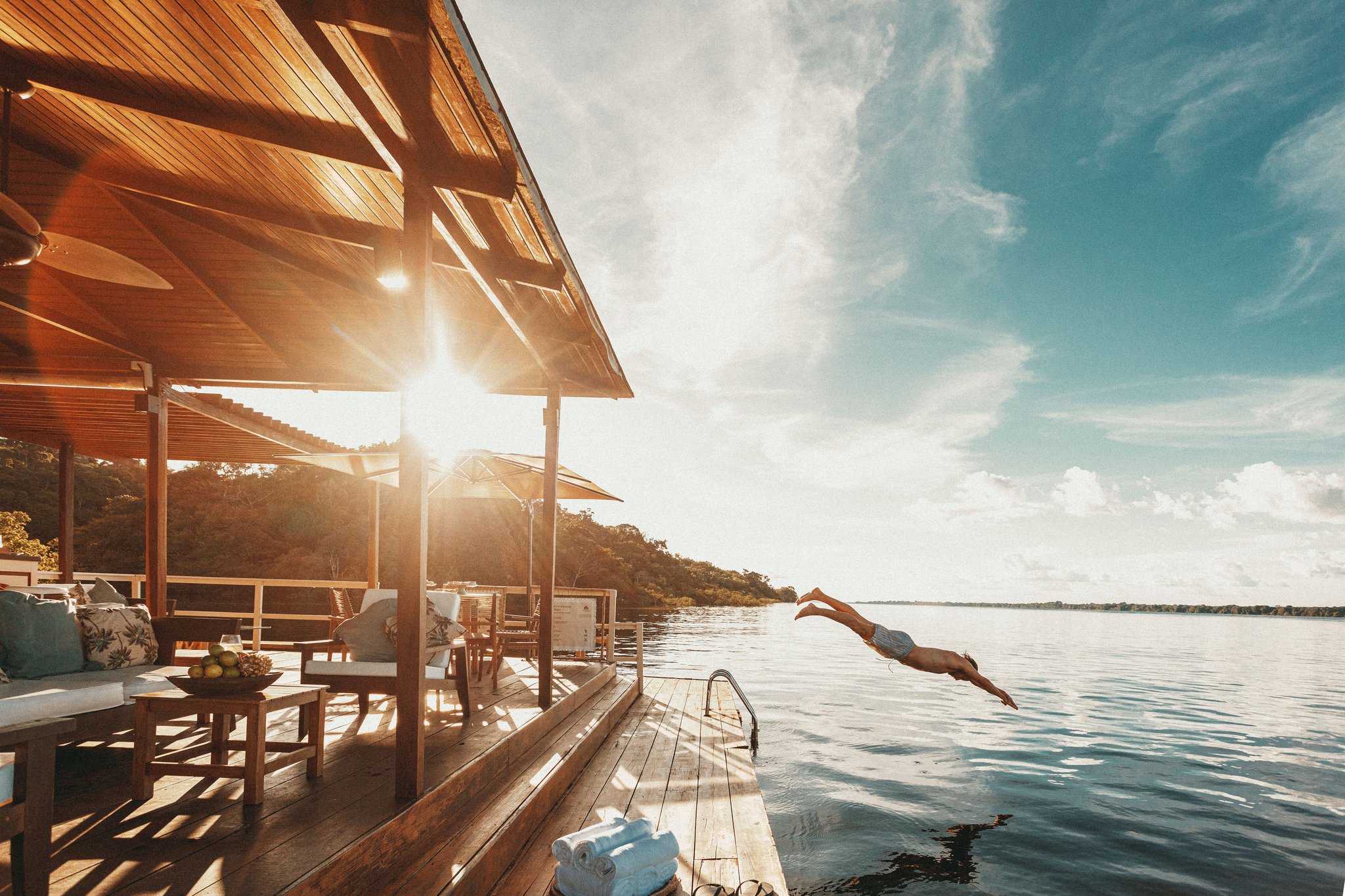 A guest dives from the wooden deck of Anavilhanas Jungle Lodge Brazil into the calm Rio Negro at golden hour, with a furnished open-air lounge and lush forest visible in the background