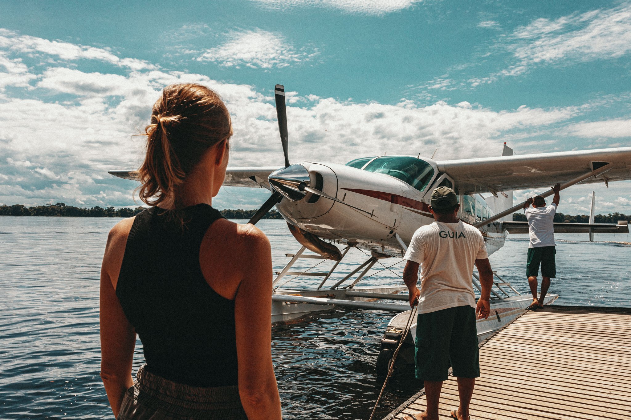 A traveller watches from a wooden dock as two guides prepare a white floatplane on the Rio Negro, ready to depart for Anavilhanas Jungle Lodge Brazil