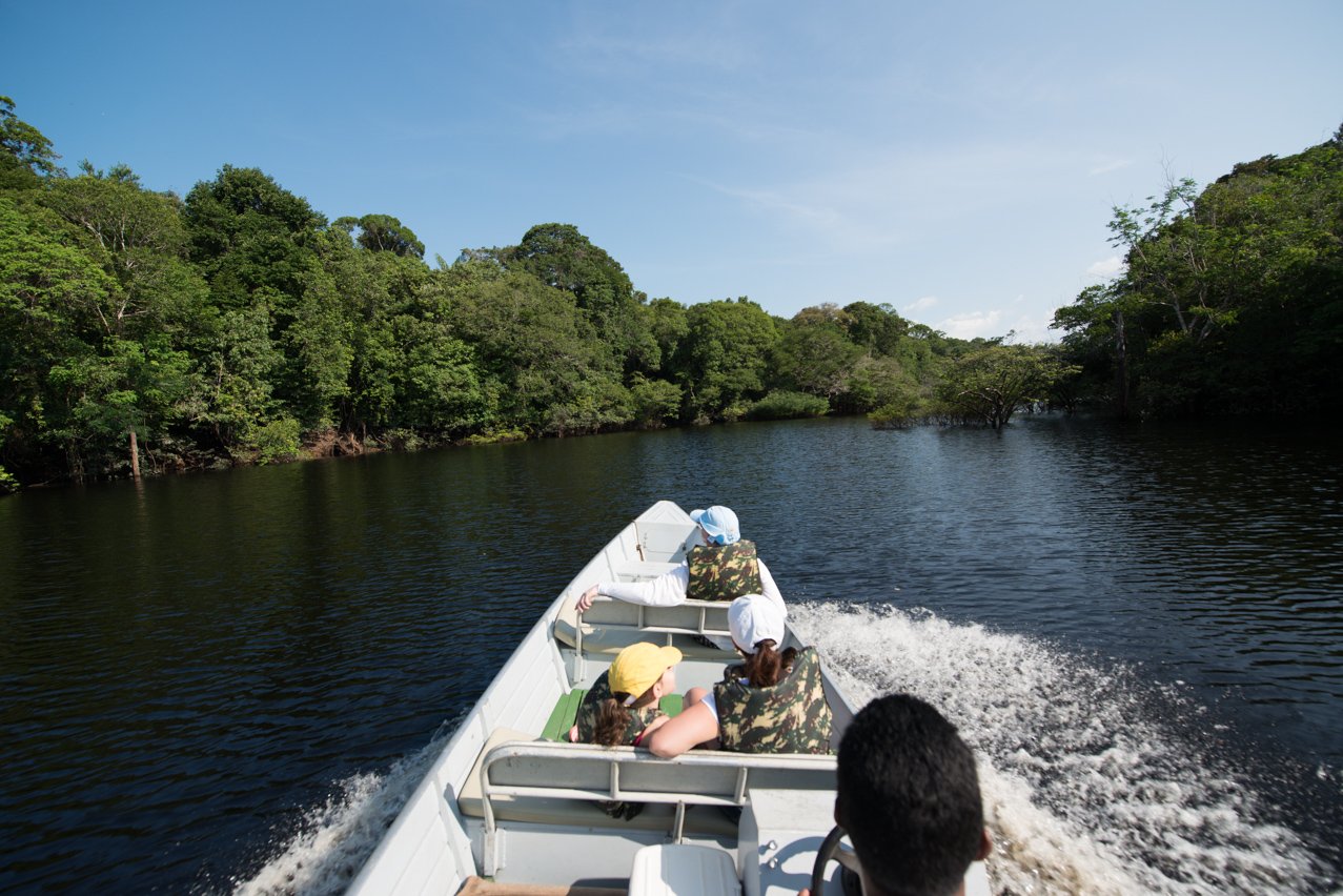 Guests wearing life jackets speed along a dark-water Amazonian river on a motorboat excursion at Anavilhanas Jungle Lodge Brazil, with dense rainforest canopy lining both banks under a clear blue sky