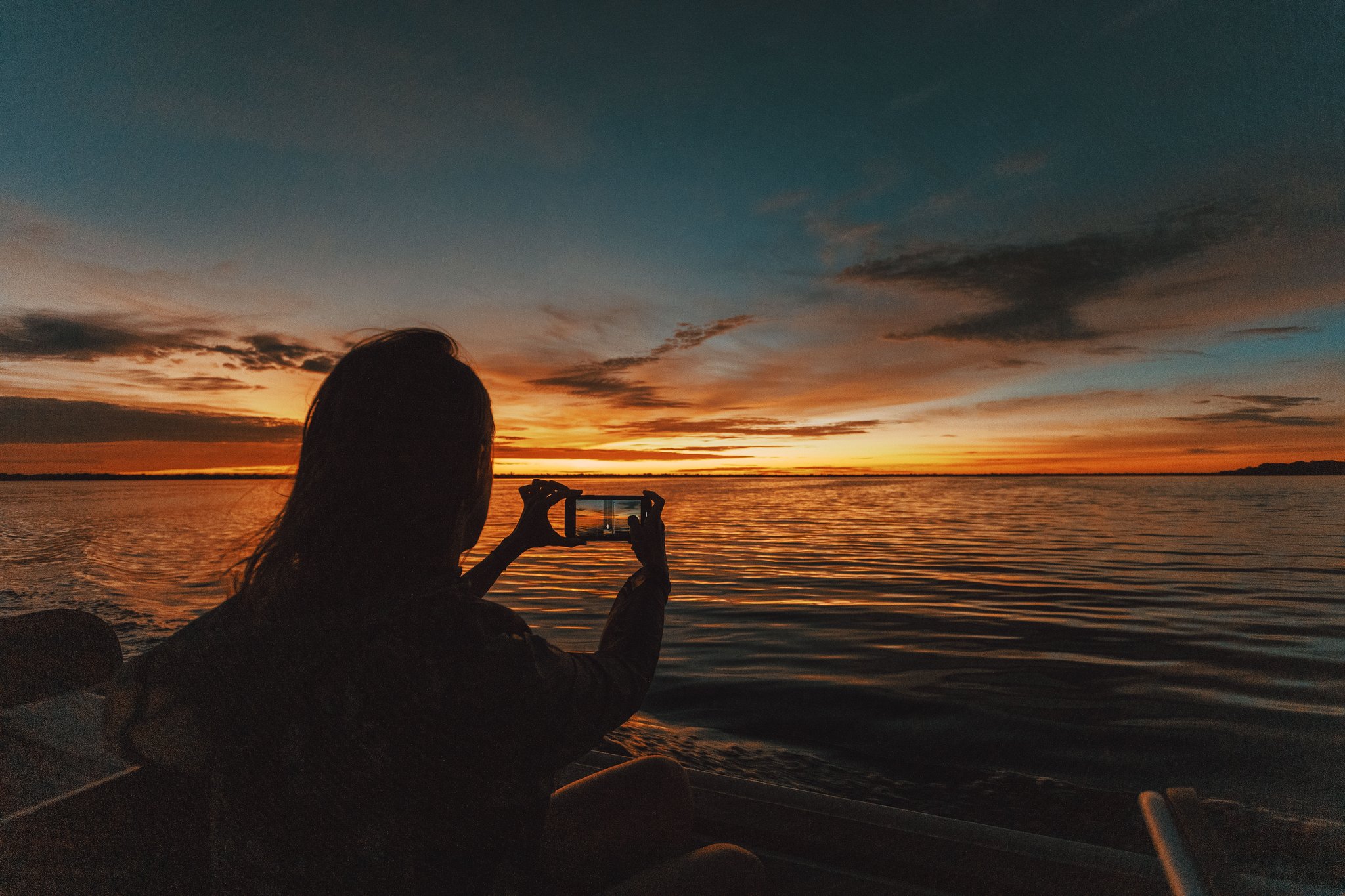 Phone raised, the whole Amazon sky caught in one small screen. A traveller silhouetted against a blazing orange and teal sunset photographs the wide river from a boat at Anavilhanas Jungle Lodge Brazil