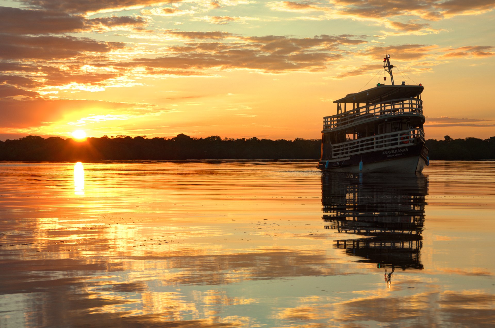 The Anavilhanas Jungle Lodge Brazil expedition boat moored on the glassy Rio Negro beneath a blazing orange sunset, its reflection perfectly mirrored on the still water
