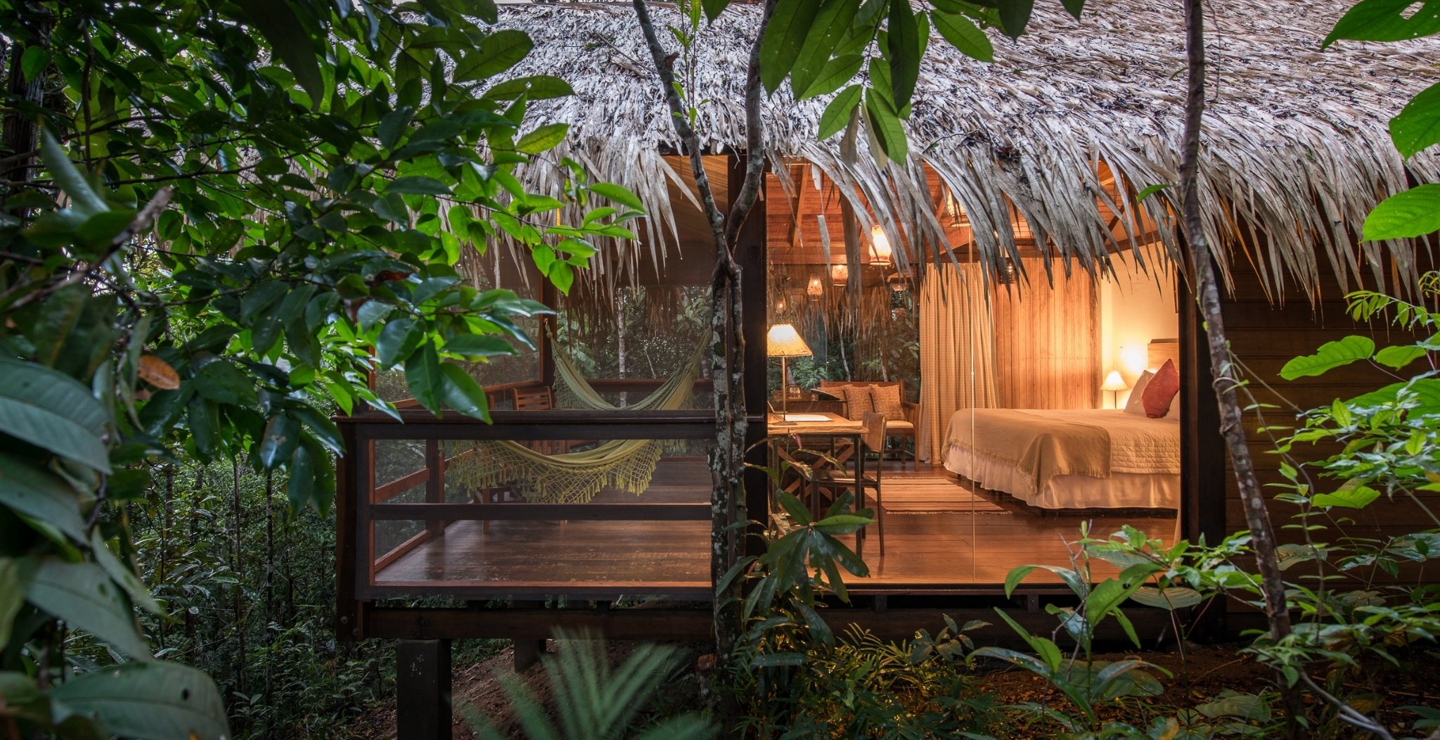 A warmly lit thatched-roof bungalow at Anavilhanas Jungle Lodge Brazil, with a woven hammock on the open deck and a king bed visible through floor-to-ceiling glass walls, surrounded by dense rainforest foliage at dusk