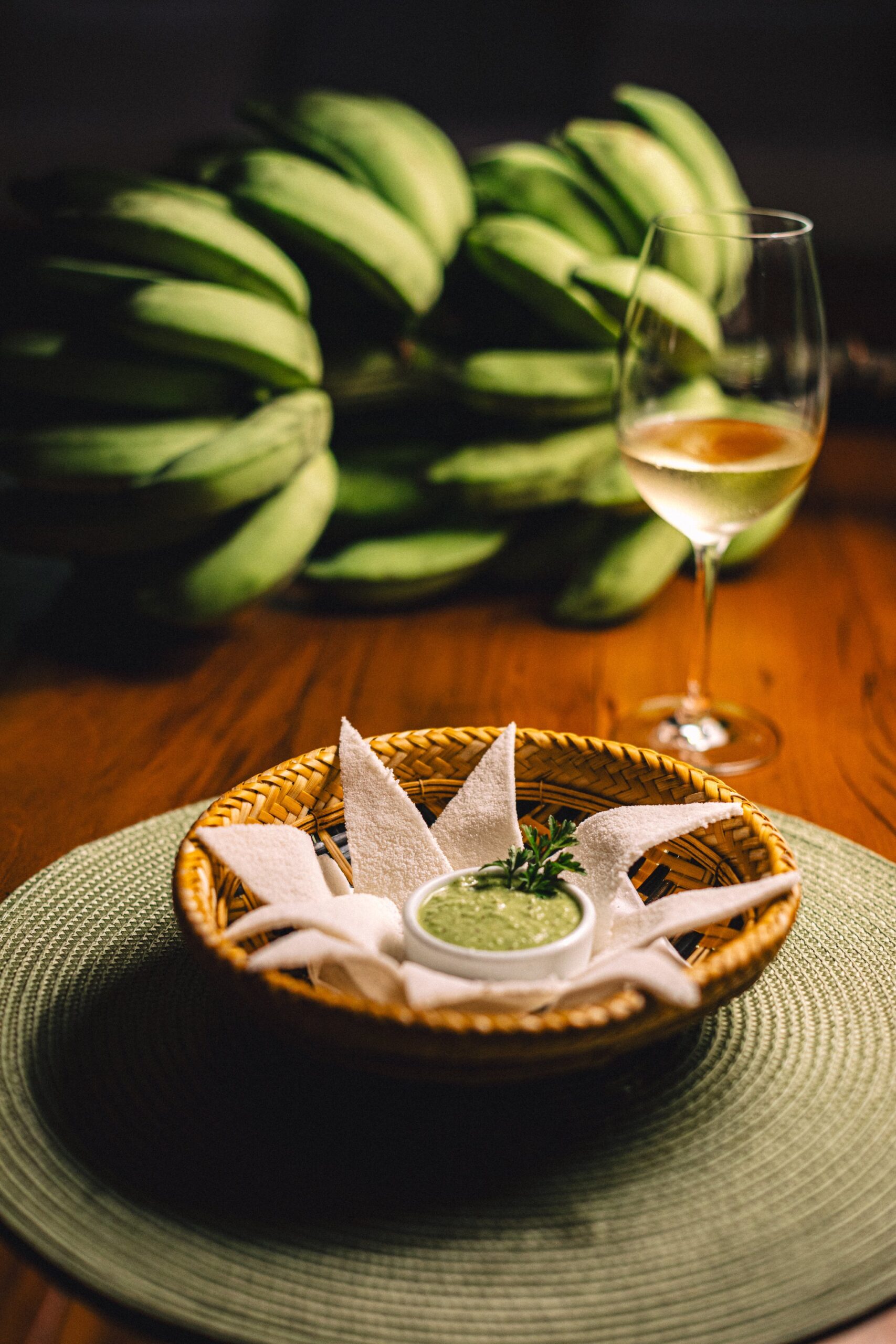 A woven rattan basket holding white flatbread pieces arranged around a small bowl of green herb dip, served at Anavilhanas Jungle Lodge Brazil alongside a glass of white wine, with a bunch of green bananas softly blurred in the background